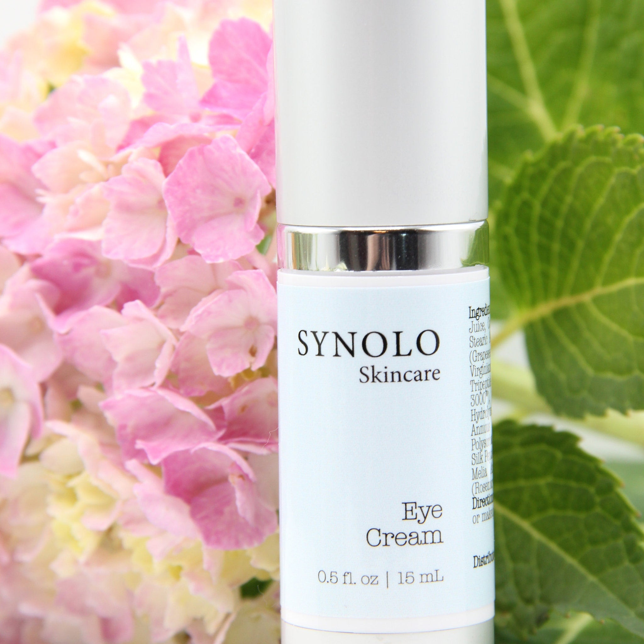 Eye cream bottle with white and silver accents, Synolo Skincare label, and pink flowers in background