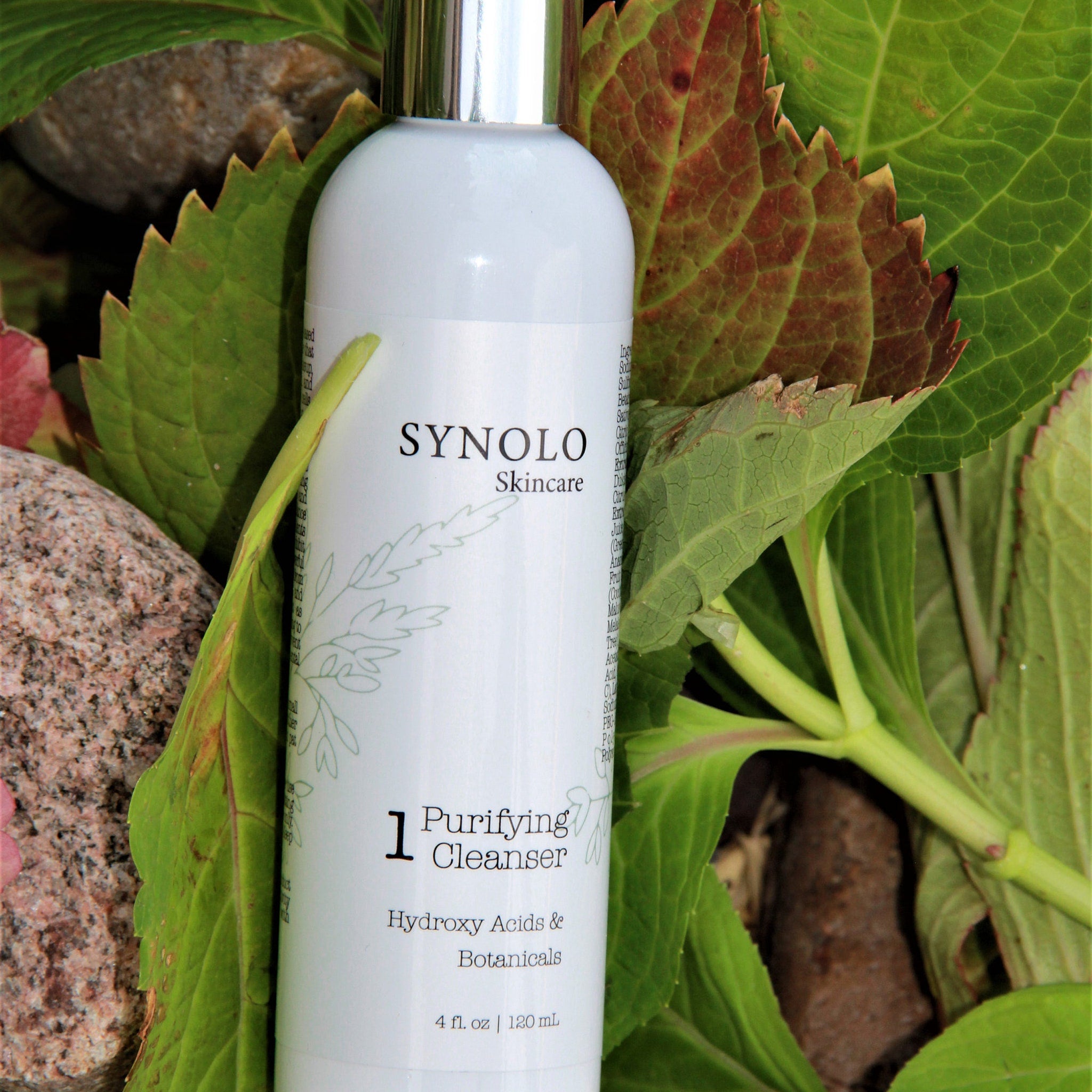 Purifying cleanser bottle, white with silver cap, surrounded by green and red leaves, Synolo Skincare visible