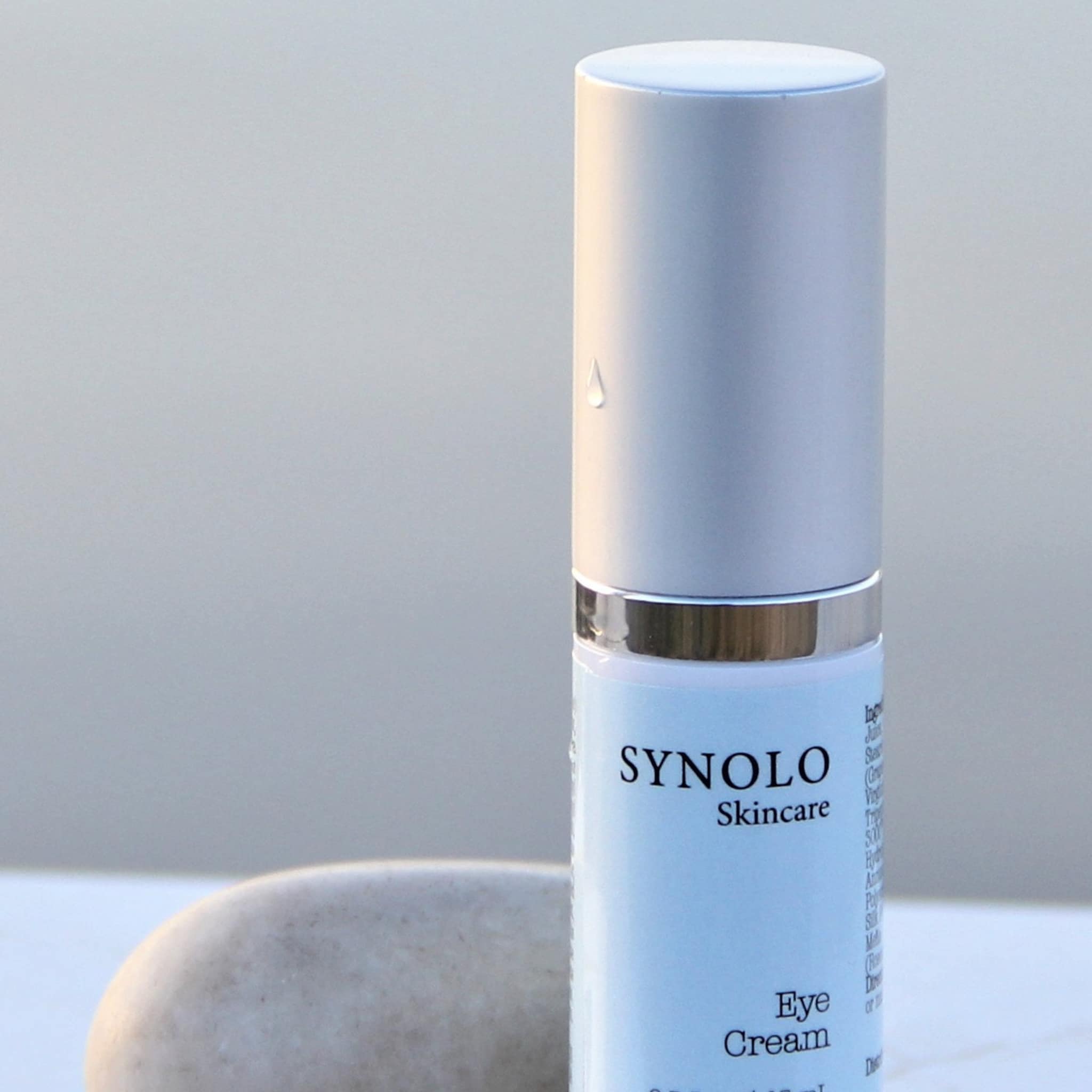 Eye cream bottle with silver and blue color, Synolo Skincare branding, and a drop detail