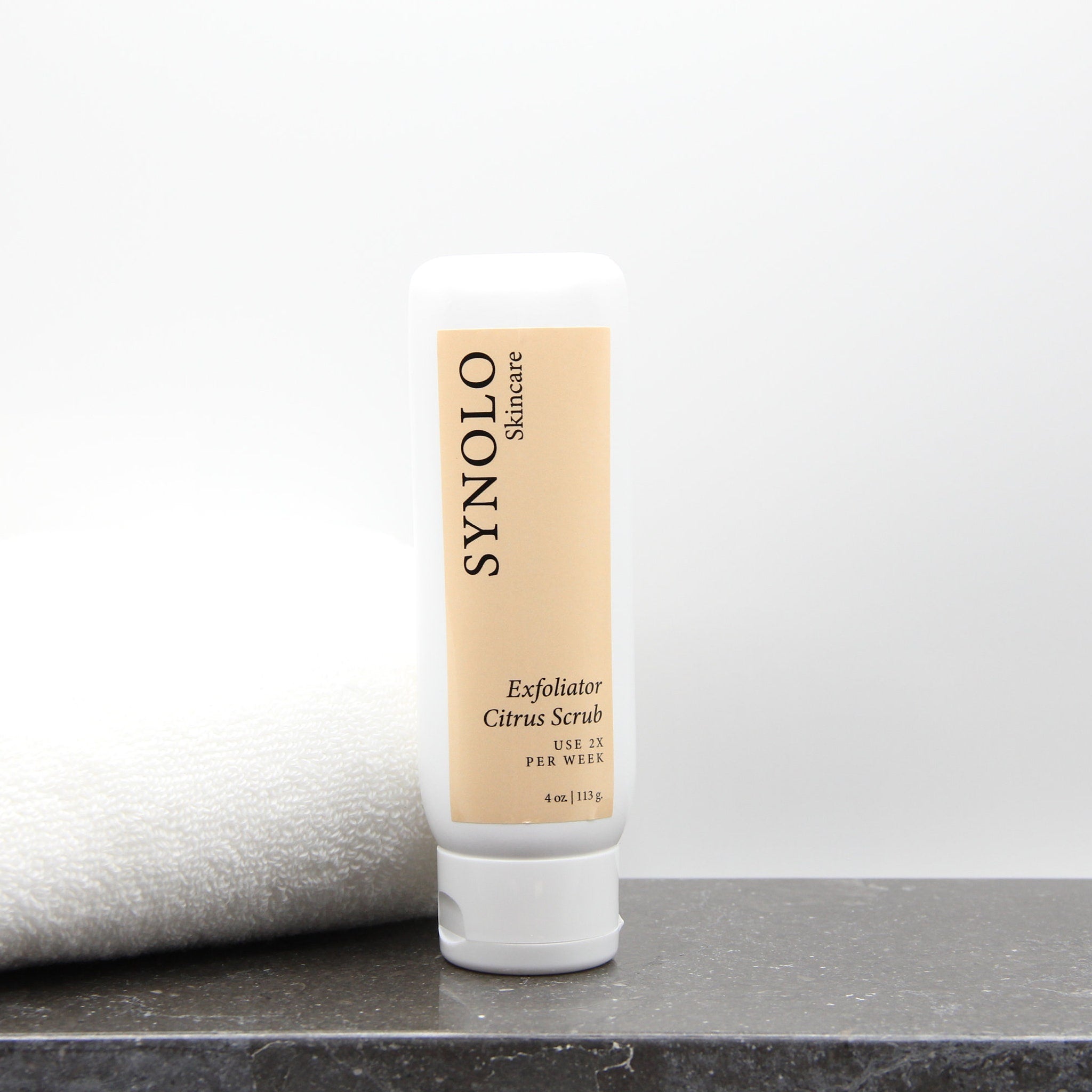 Exfoliator Citrus Scrub white bottle with Synolo Skincare label on a counter next to a towel