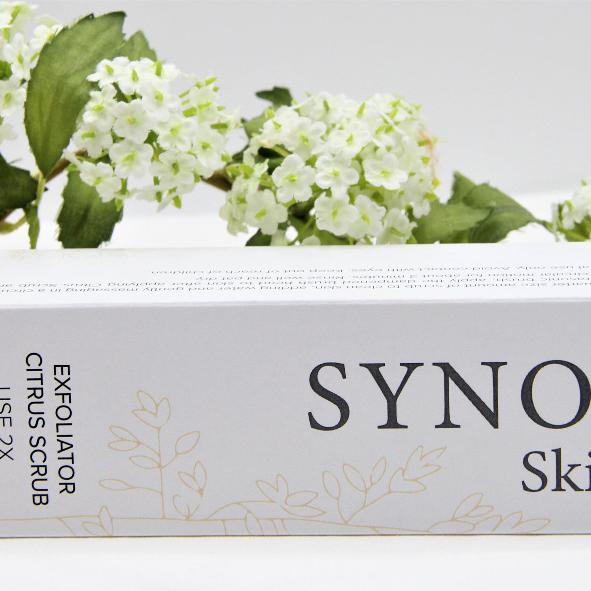 Exfoliator Citrus Scrub box with white and gold floral design, "SYNO" text, and white background