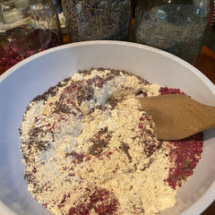 Mixture of beige powder, red petals, and brown herbs in a white bowl with wooden spoon