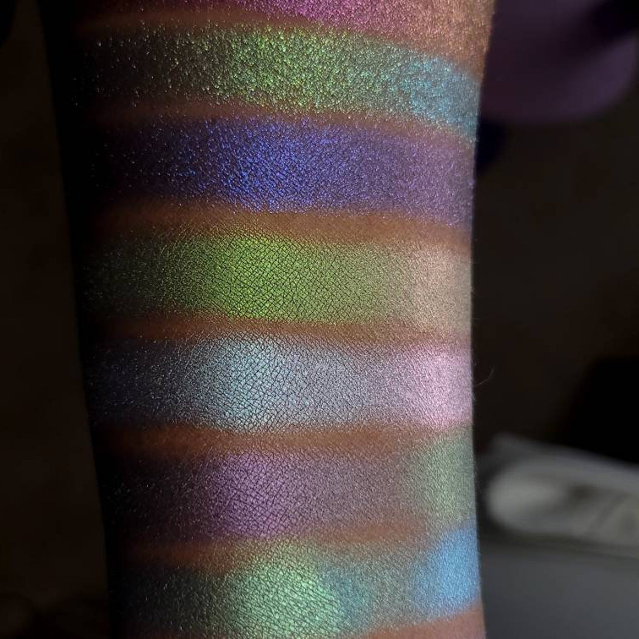 Eyeshadow swatches with iridescent green, purple, and blue hues on arm, showing color variations