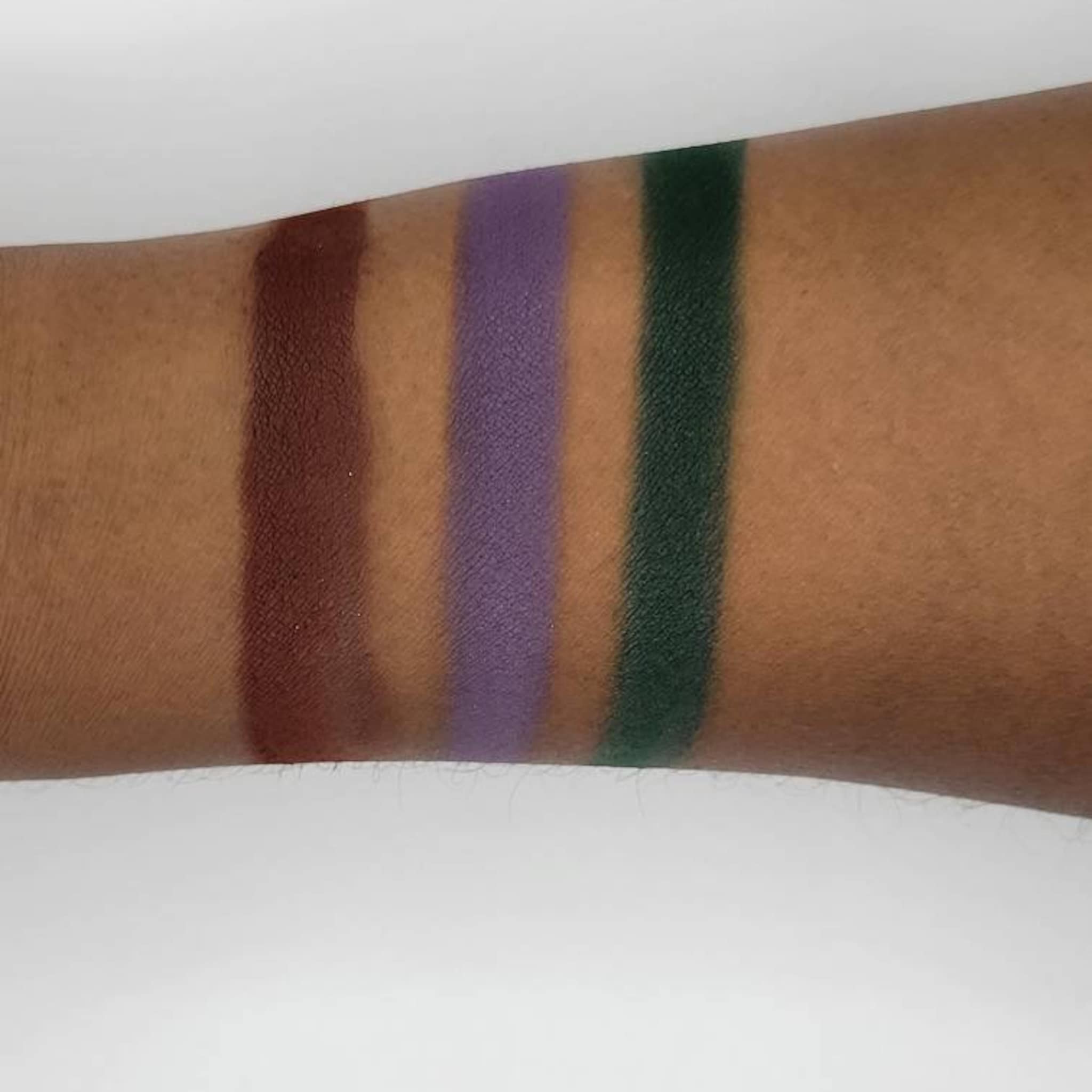 Eyeshadow swatches in brown, purple, and green shades on arm, showing color and texture