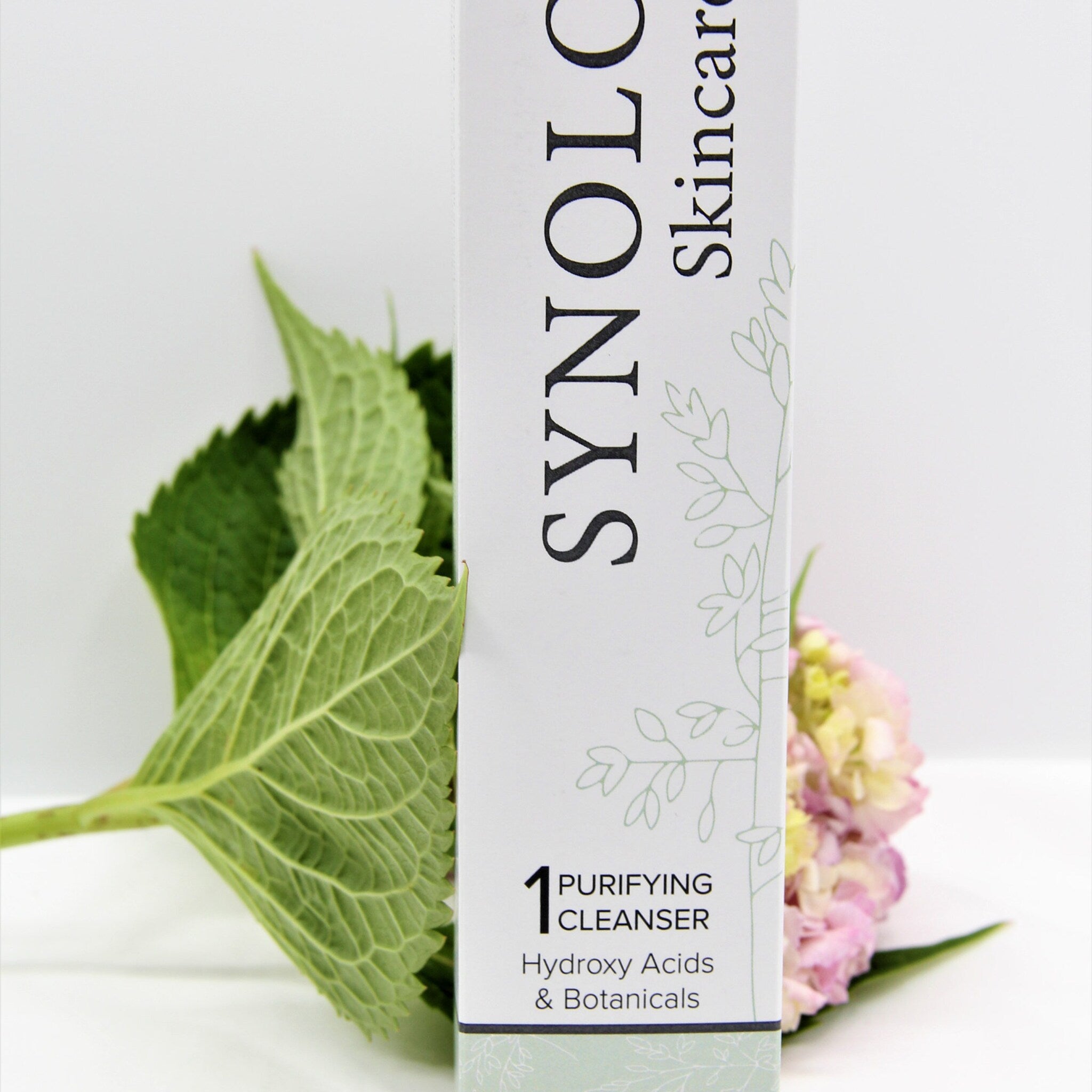 Purifying cleanser box with white and green floral design, green leaves, and pink flowers in the background