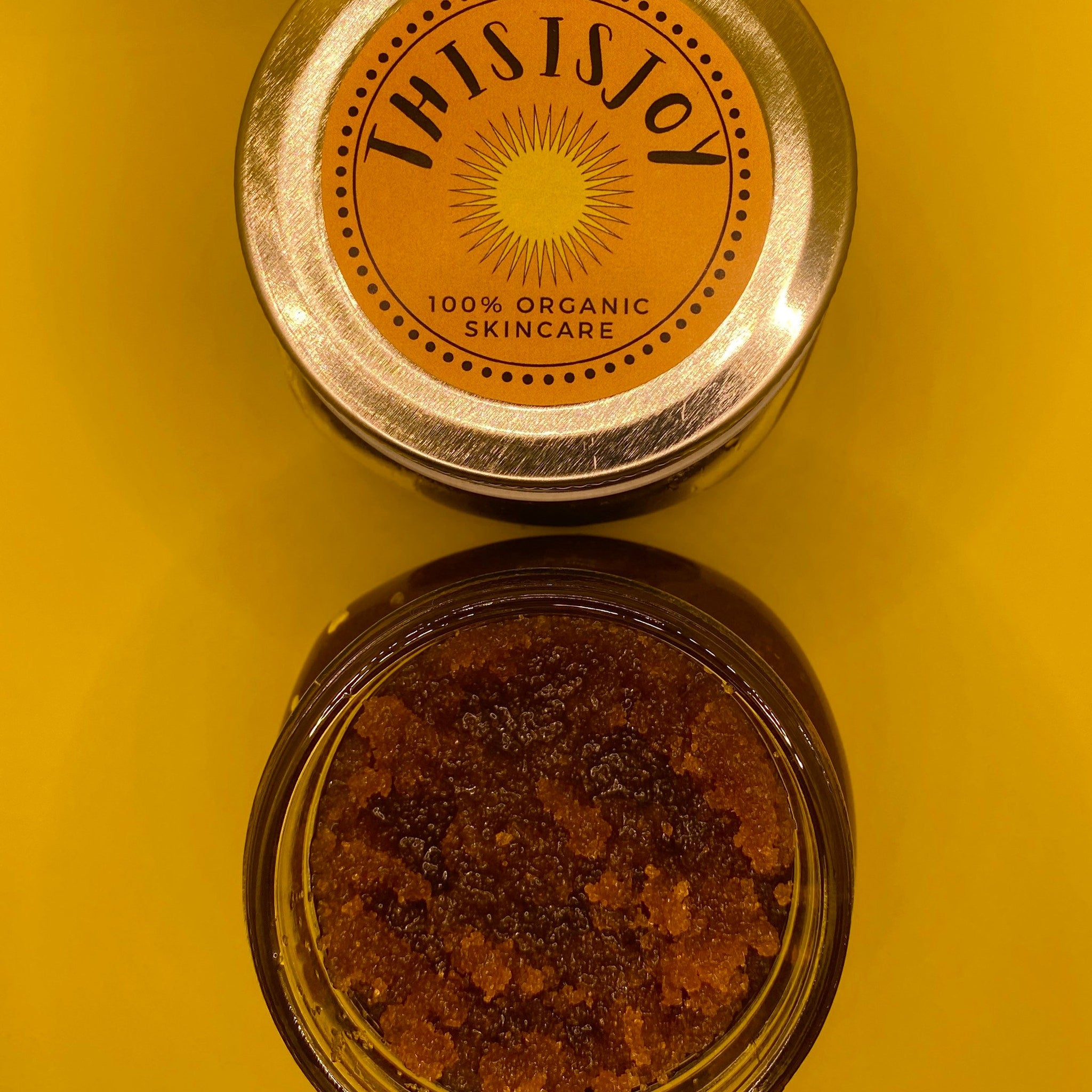 Skincare jar with brown granules, gold lid, and "THIS IS JOY" label on yellow background