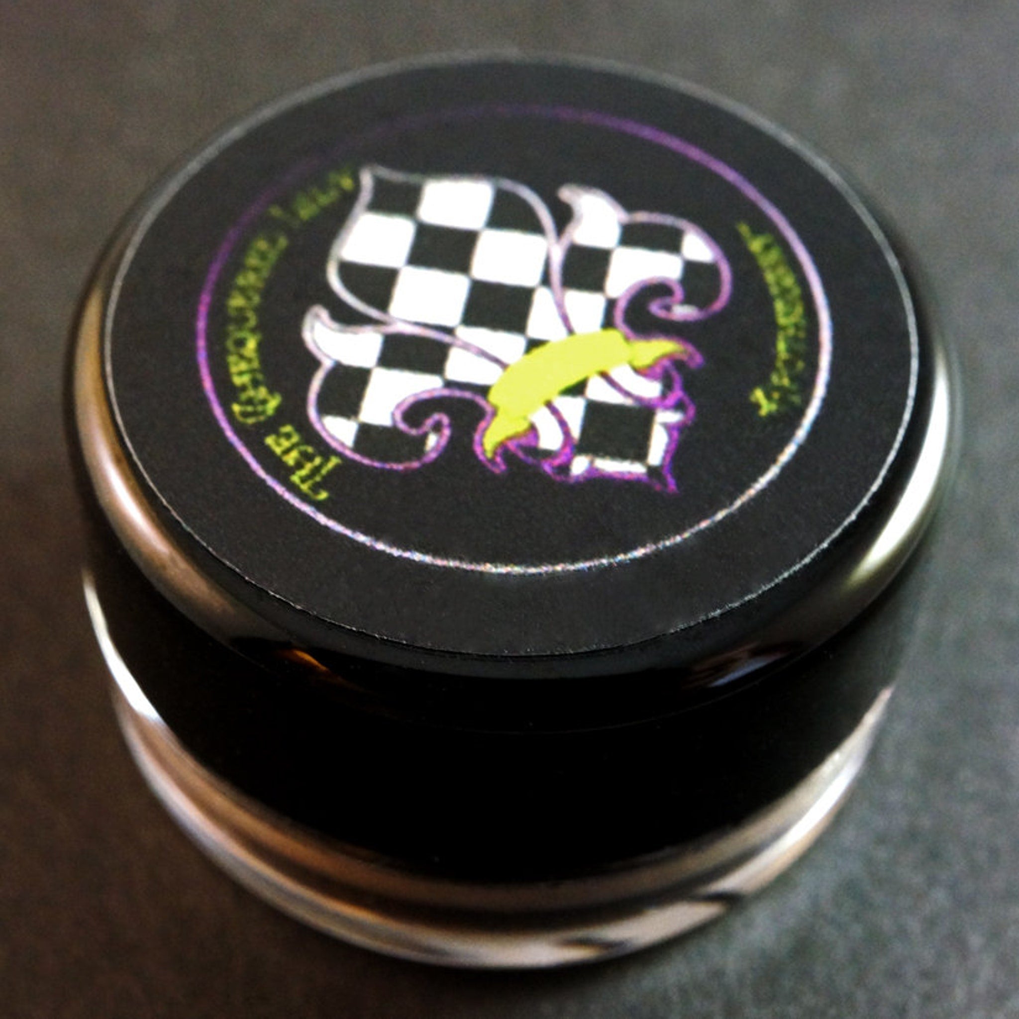 Black round container with checkered pattern and "The Key Sequence" text, reflective rim