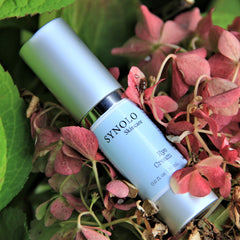Eye cream bottle with silver cap, white label, and SYNOLO Skincare text, surrounded by pink flowers
