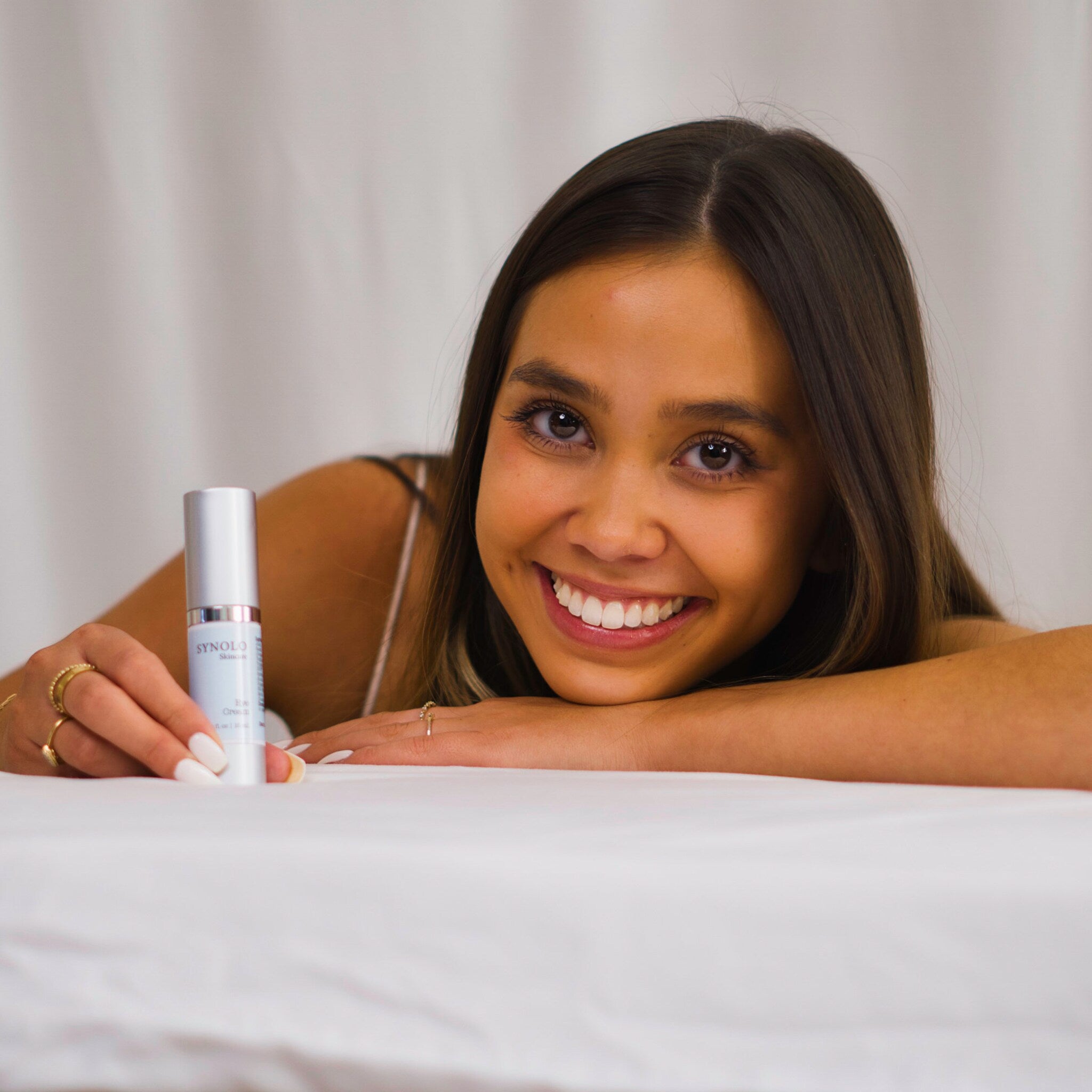 Eye cream bottle, silver and white, held by woman with long brown hair, Synolo Skincare label visible