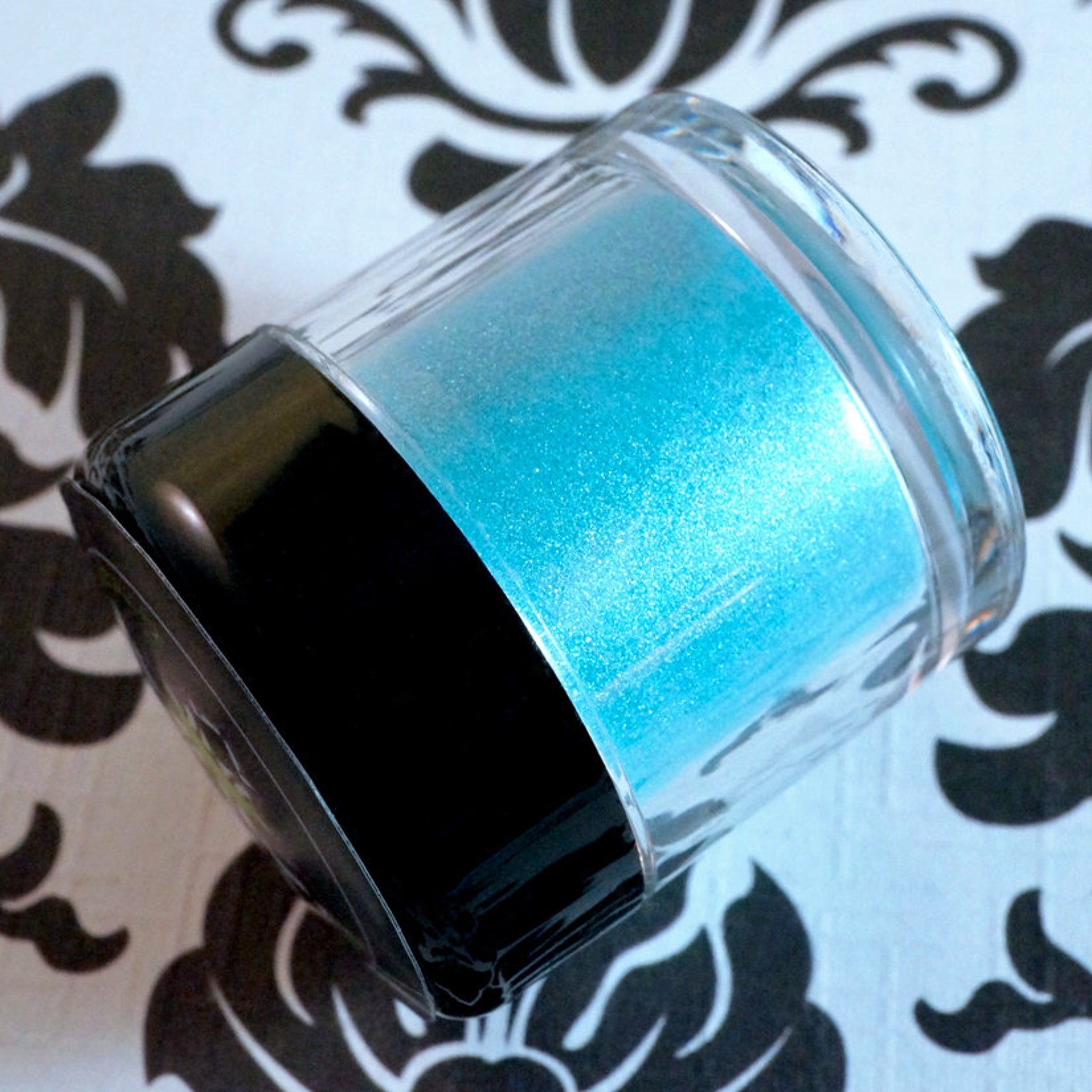 Swatch of iridescent teal and purple loose eye shadow with shimmer on fingertip against neutral background