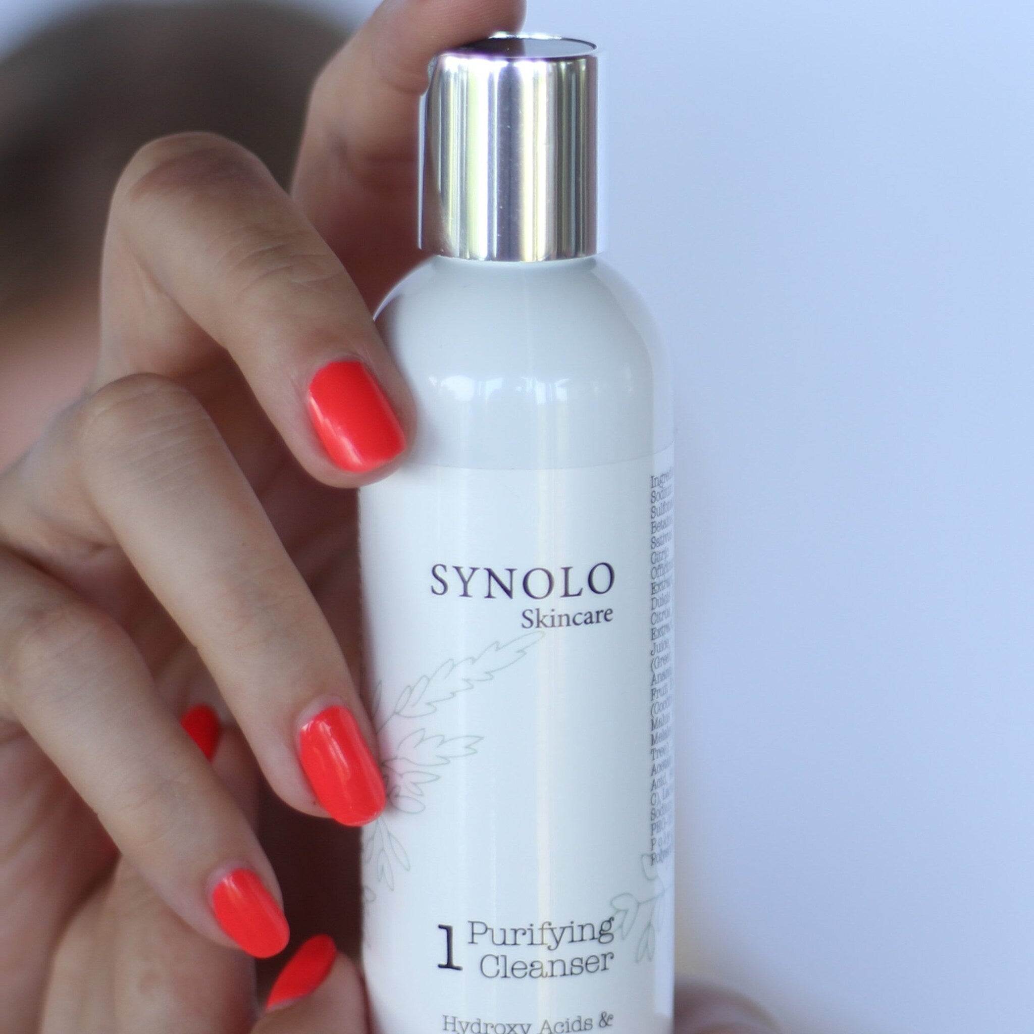 Purifying cleanser bottle, white with silver cap, held by hand with red nails