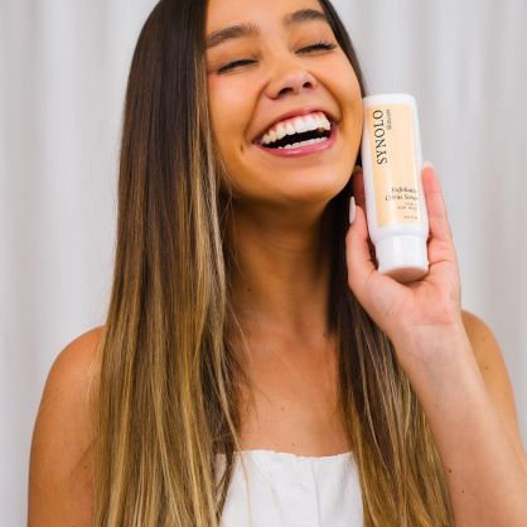 Skincare product bottle, white and beige, held by smiling woman with long hair, SYNOLO brand visible