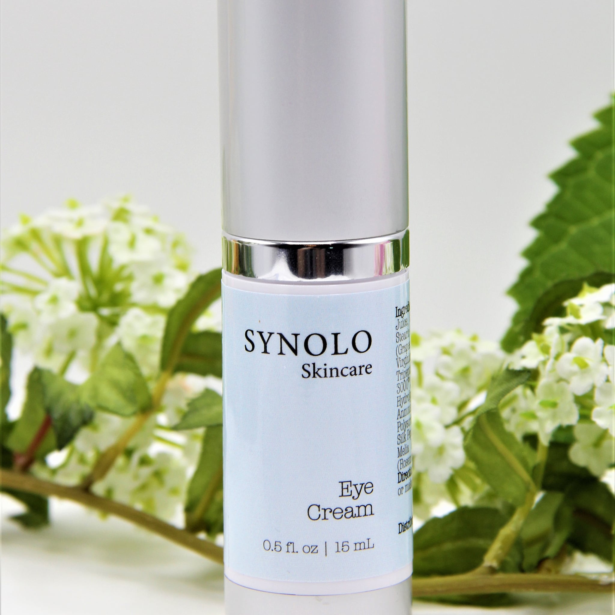 Eye cream bottle with silver cap, light blue label, and text "SYNOLO Skincare" with white flowers in background