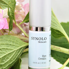 Eye cream bottle with silver cap and light blue label, SYNOLO Skincare brand, with green leaves