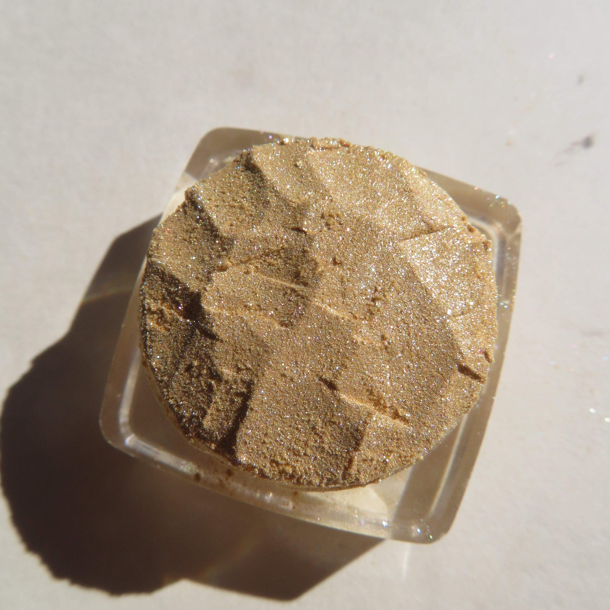 Eye shadow, gold glittery powder in clear square container, textured surface, top view