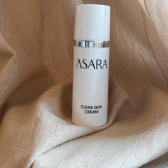 ASARA clear skin cream in white bottle with silver band on beige fabric background