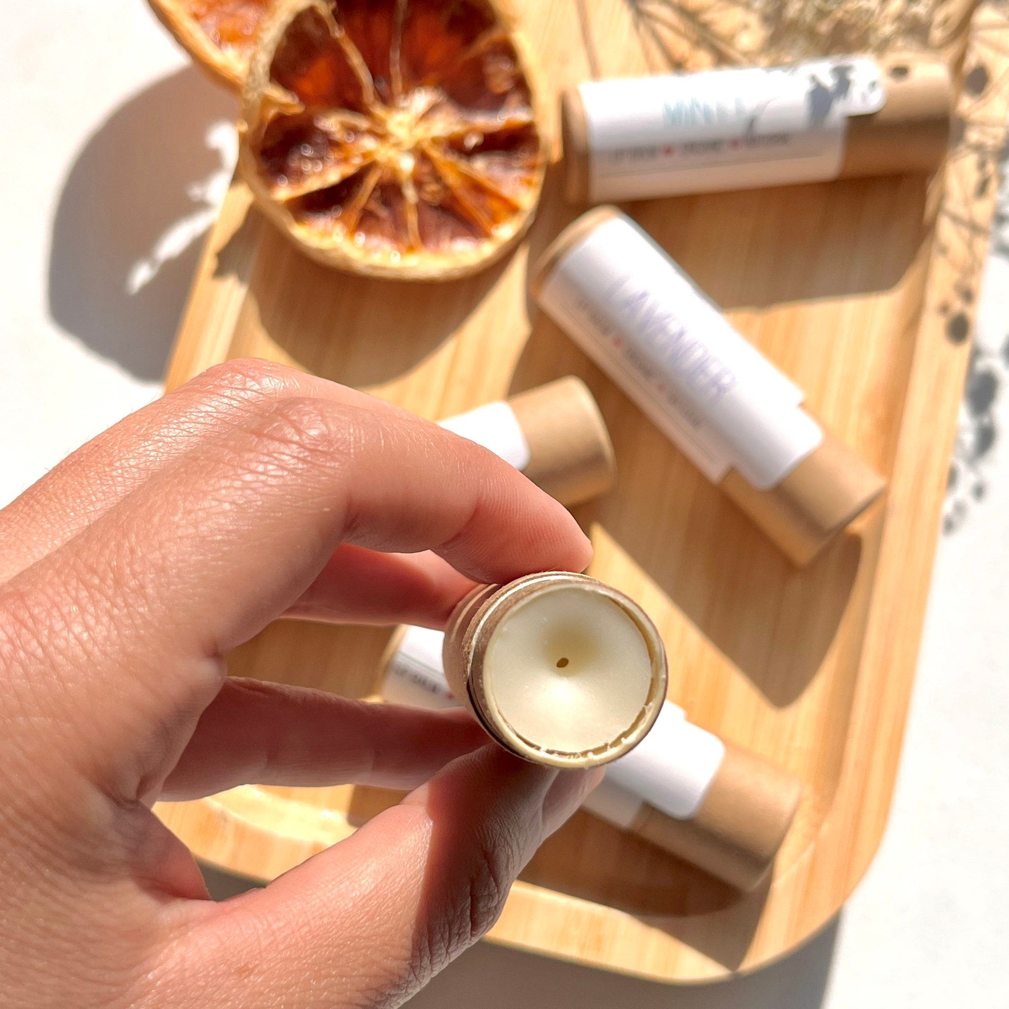 Lip balm tubes, brown paper, white label, and cream-colored balm, held by a hand on a wooden tray.