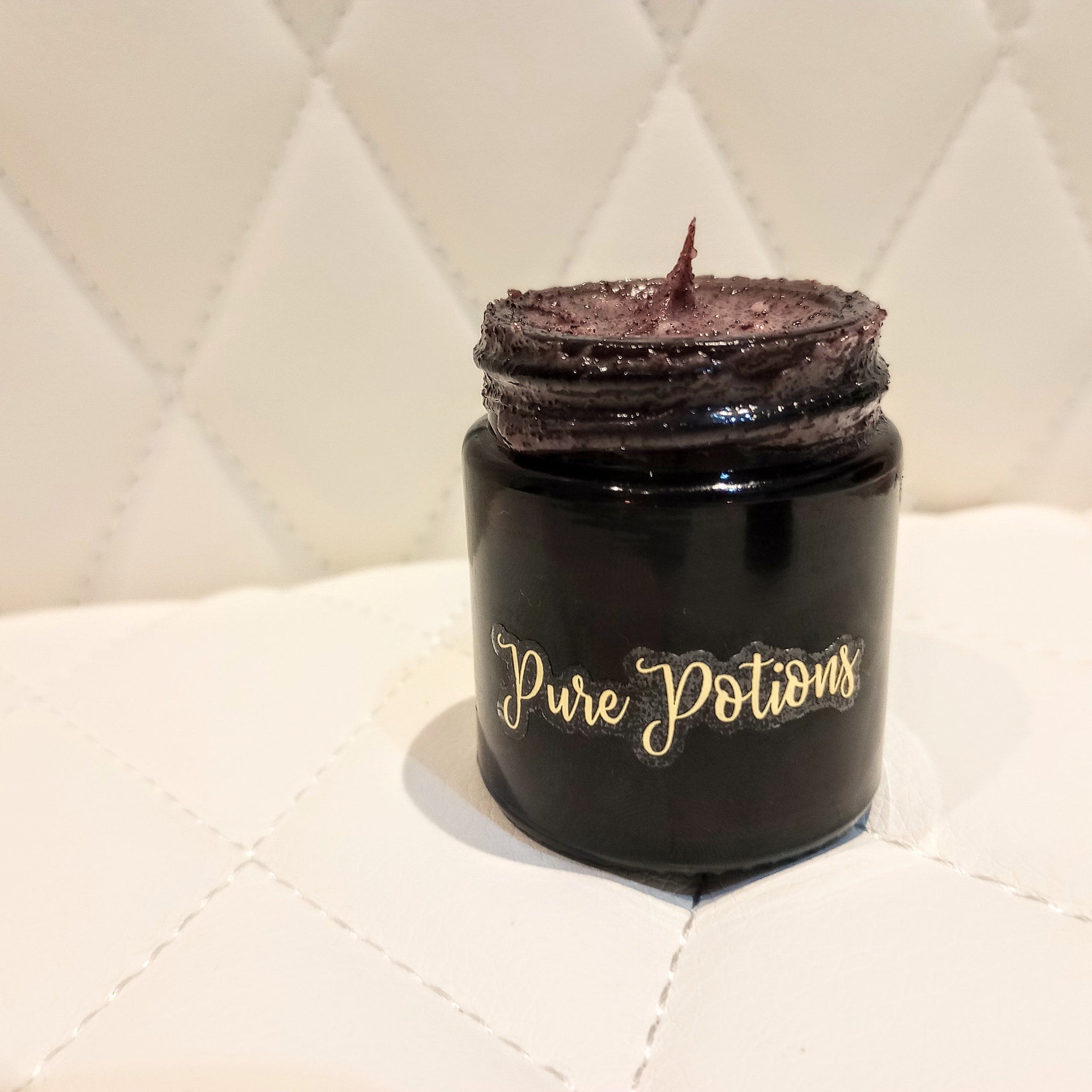 Black jar with dark purple contents, gold "Pure Potions" label, on white quilted surface
