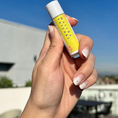 Lip balm tube with yellow label, white cap, and "Matcha Taiyaki" text held in hand against blue sky