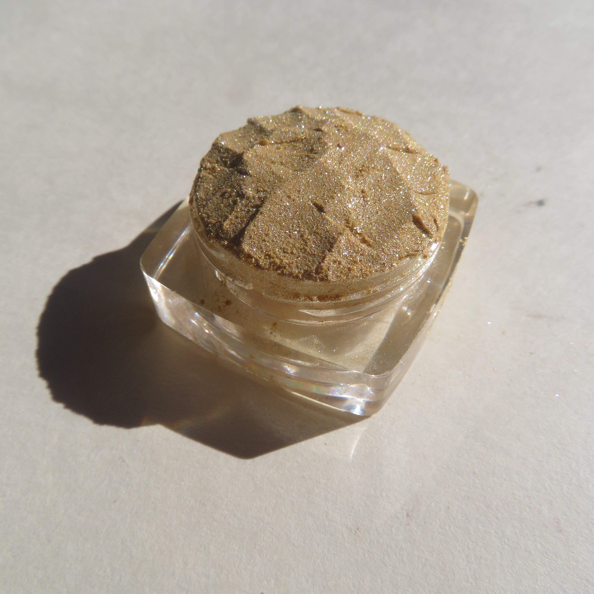 Eyeshadow pot with tan powder, clear plastic container, and a textured surface.