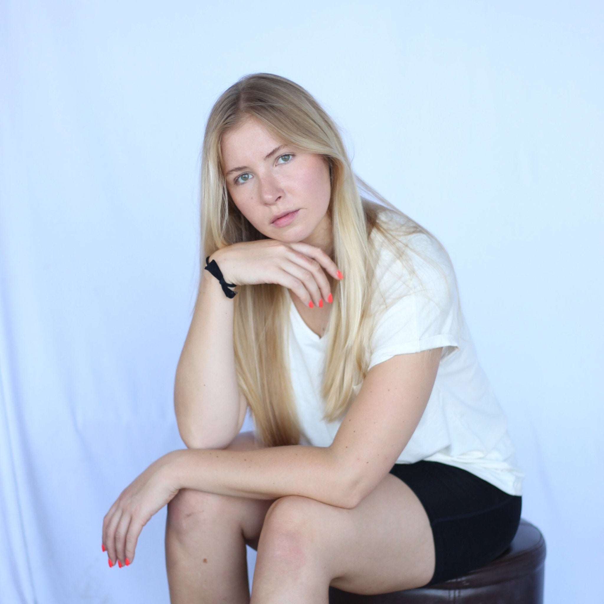 Woman with blonde hair wearing white t-shirt and black shorts, sitting on a stool, looking at the camera