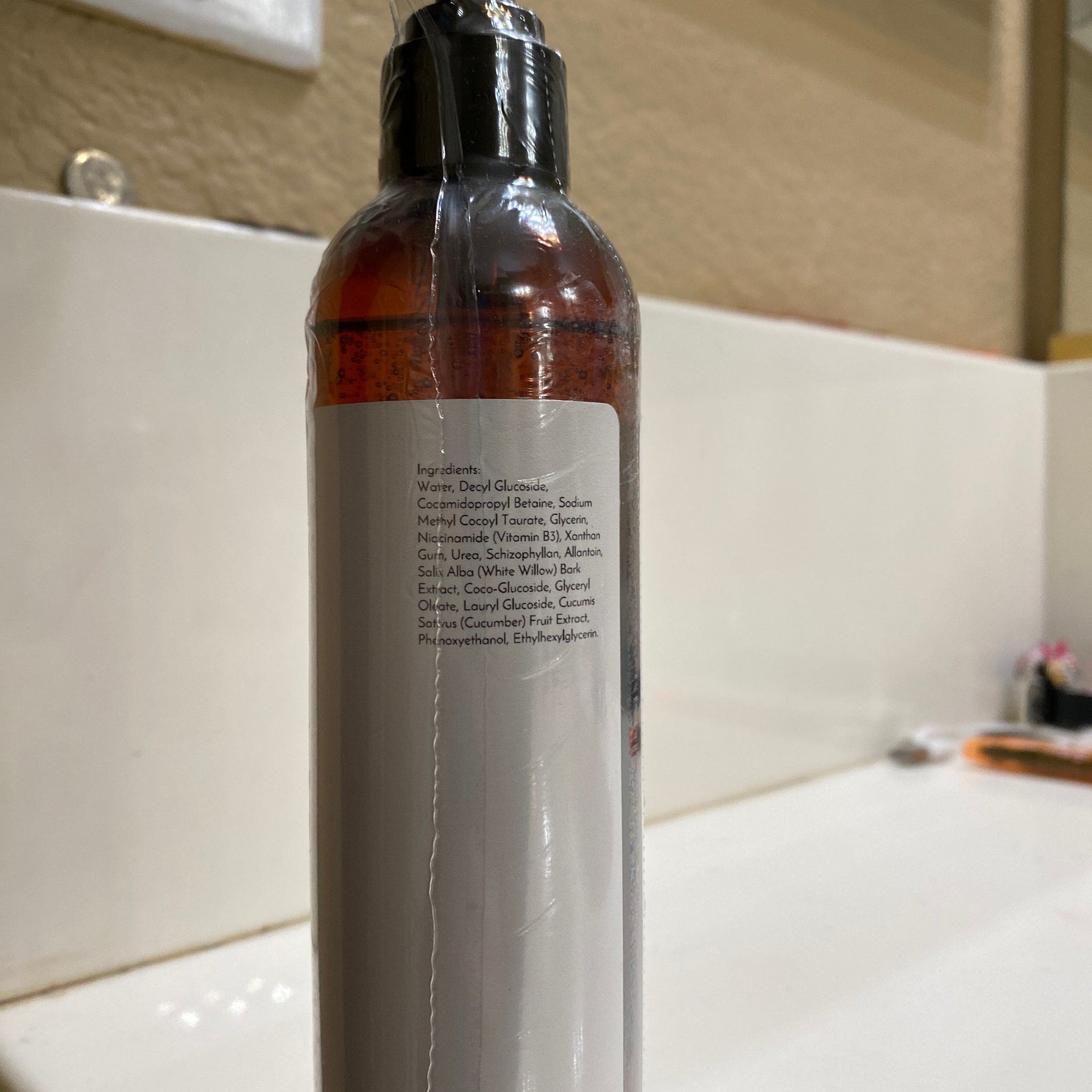 Bottle of brown liquid with white label and black cap, wrapped in clear plastic