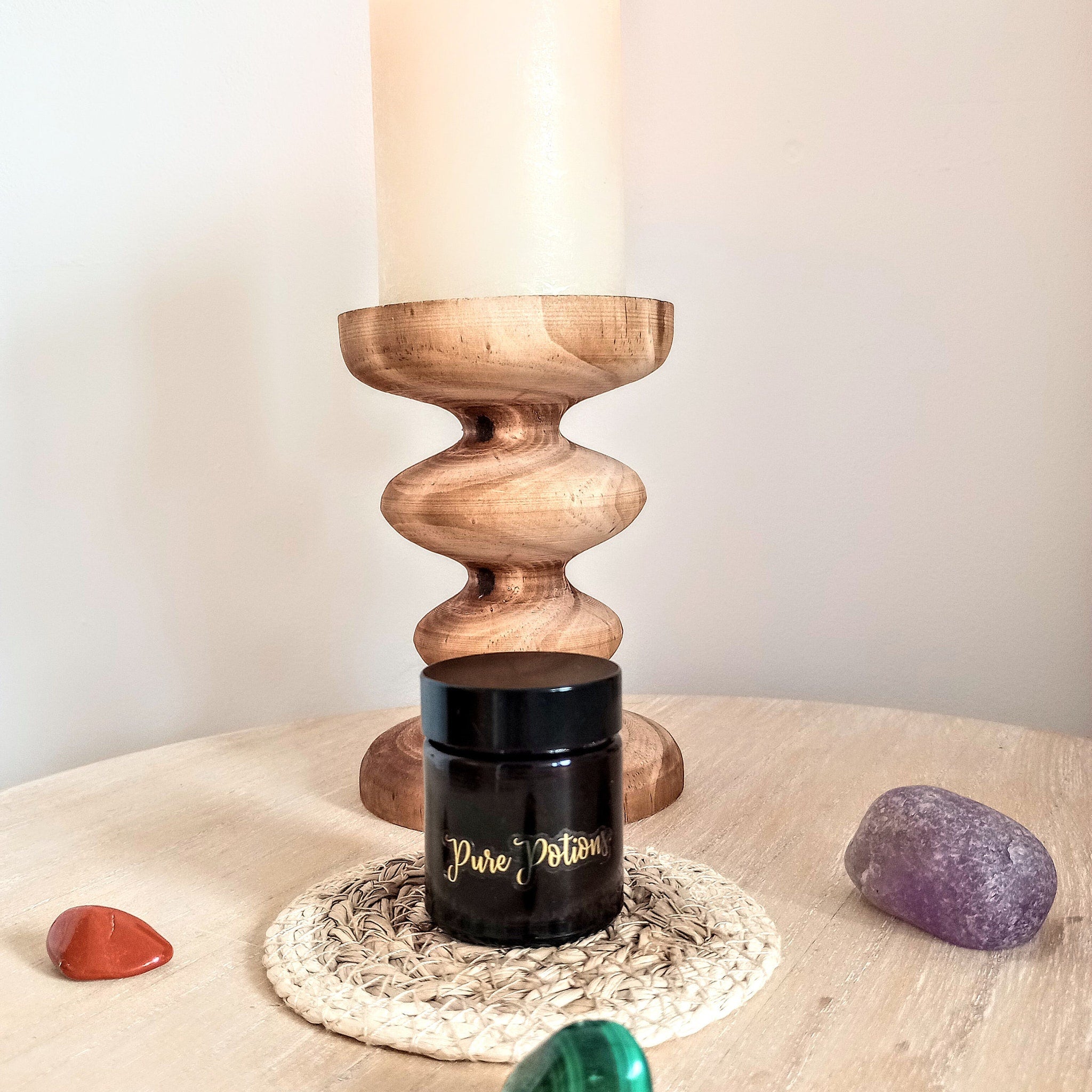 Black jar with gold "Pure Potions" text on a woven coaster, next to a wooden candle holder