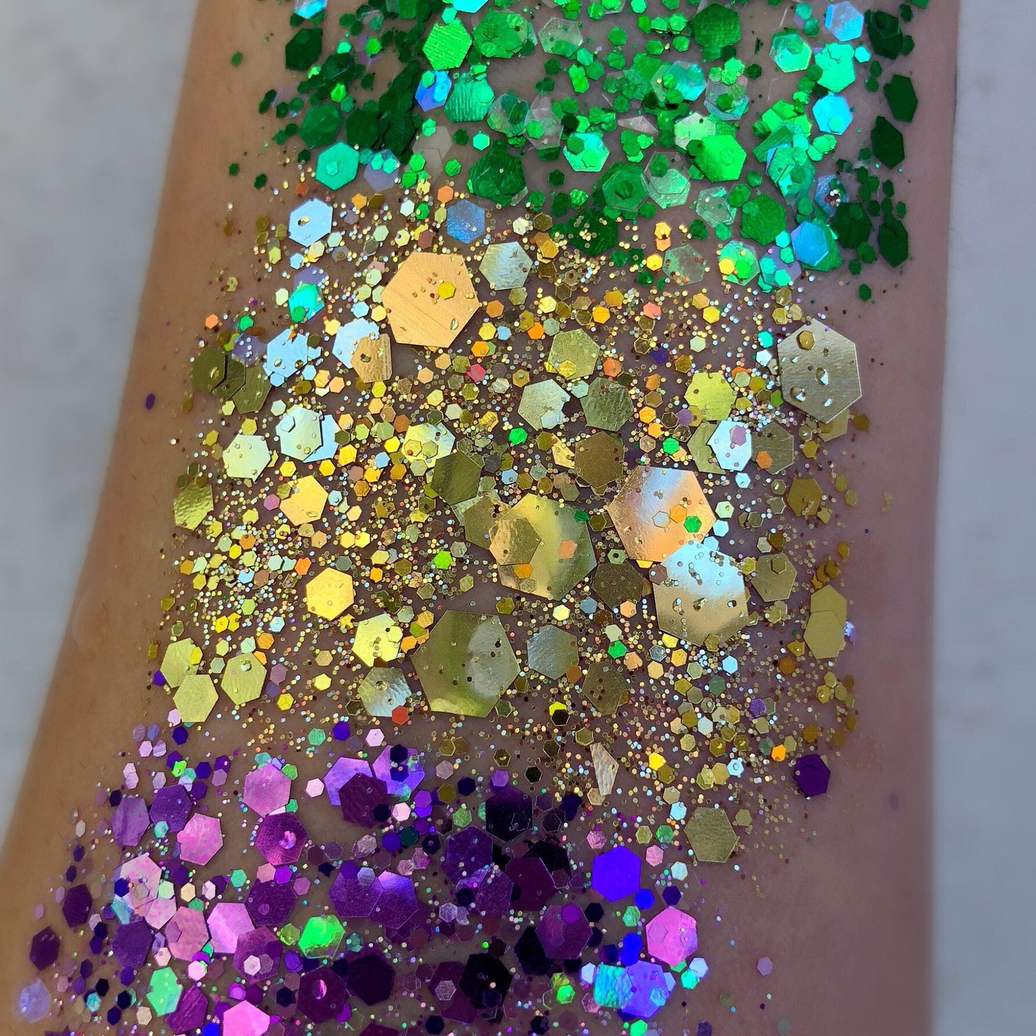 Side view of three chunky loose glitters in purple, green, and gold with adhesive bottle on marble surface