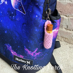 Pink crochet lip balm holder with brown lip balm attached to a blue galaxy patterned backpack