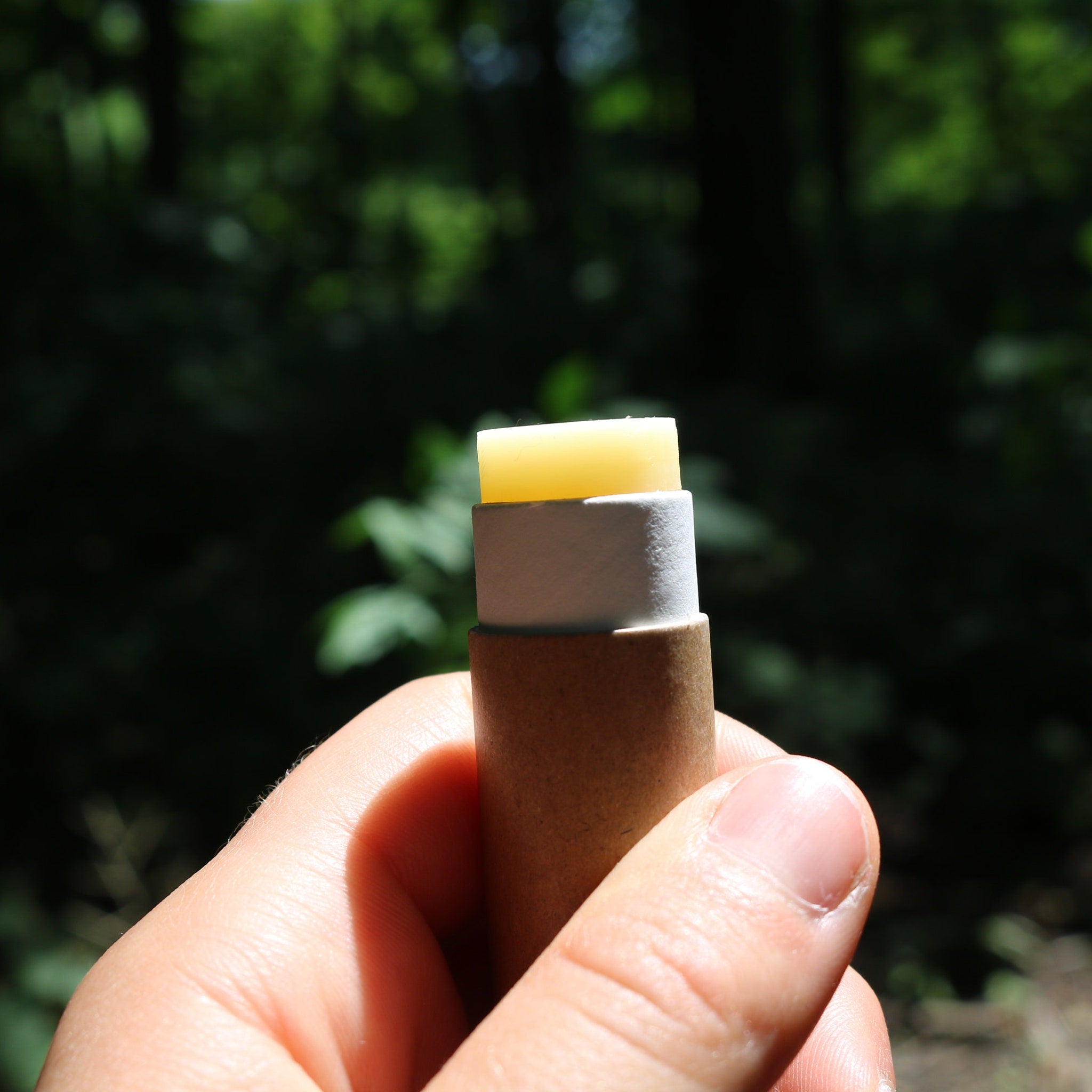 Lip balm stick with yellow, white, and brown components held in hand against a blurred green background