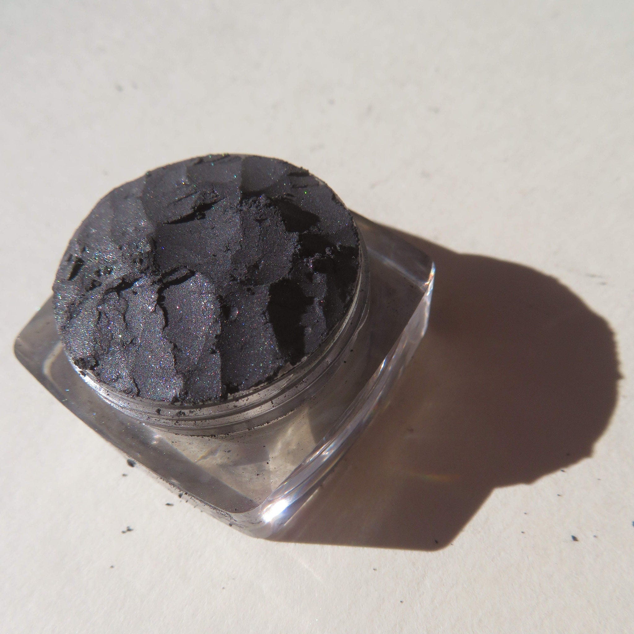 Black shimmer mineral eyeshadow in clear jar with silver lid, loose pigment spilled on marble surface