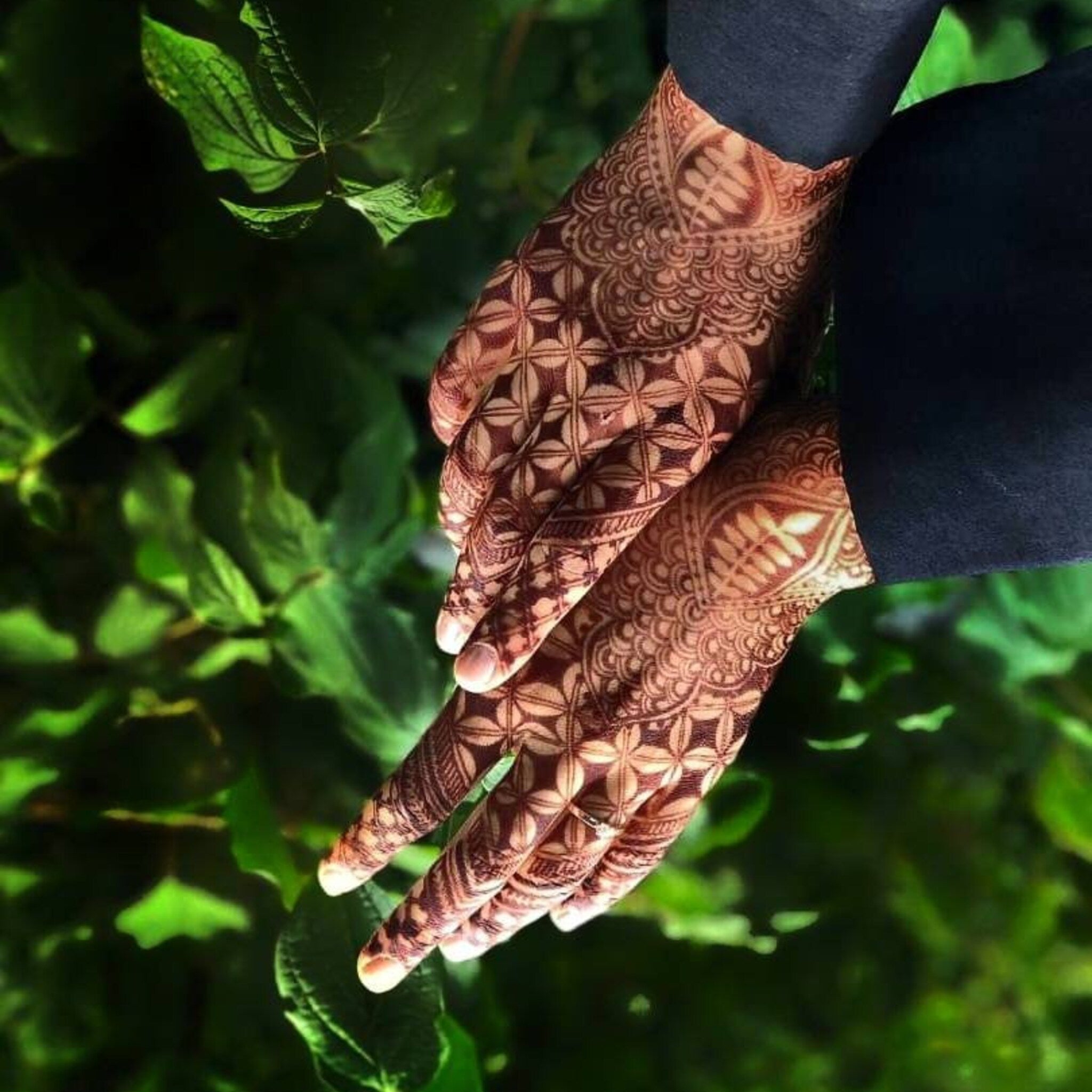 Henna hand art with brown floral pattern on skin and black sleeve against green foliage