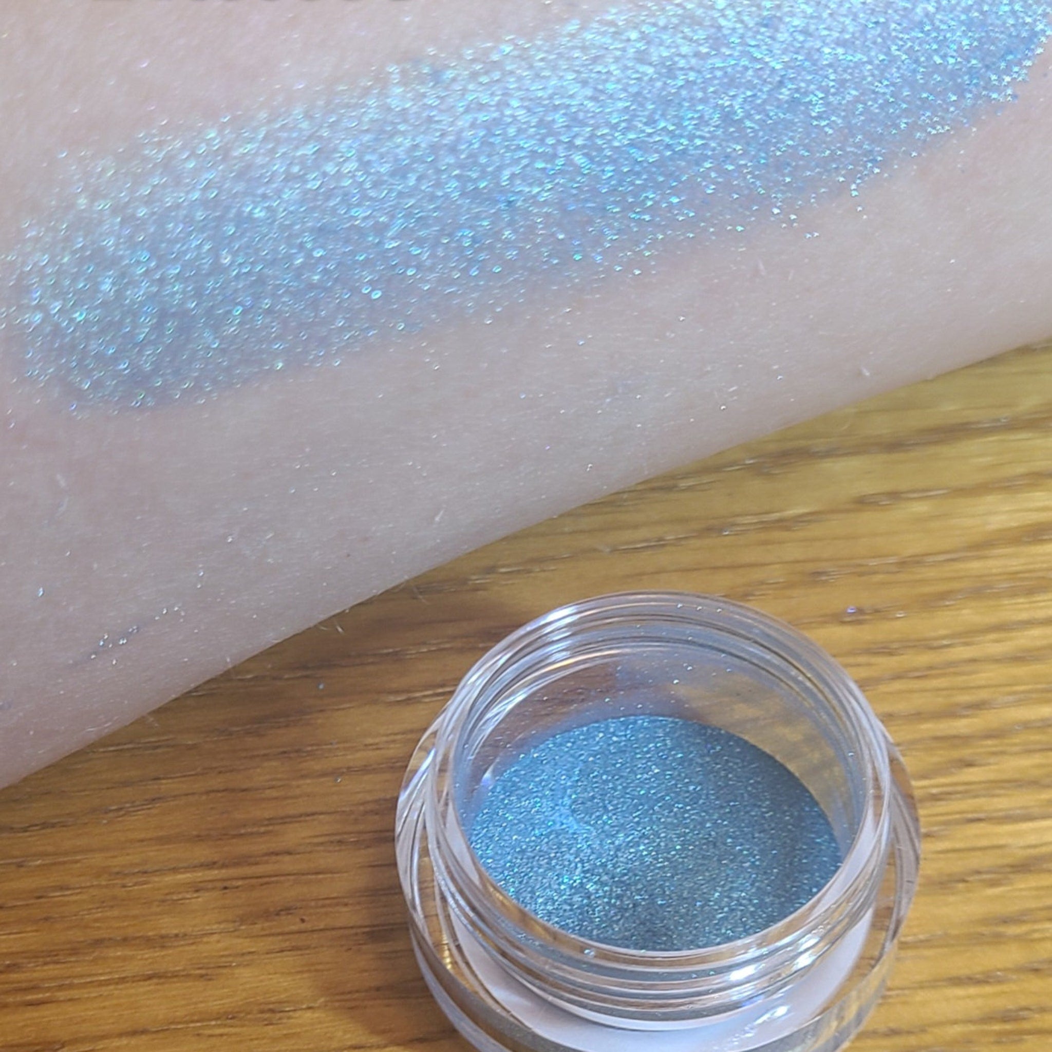 Side view of loose blue eyeshadow powder spilling from a small jar, showcasing its vibrant color and fine texture