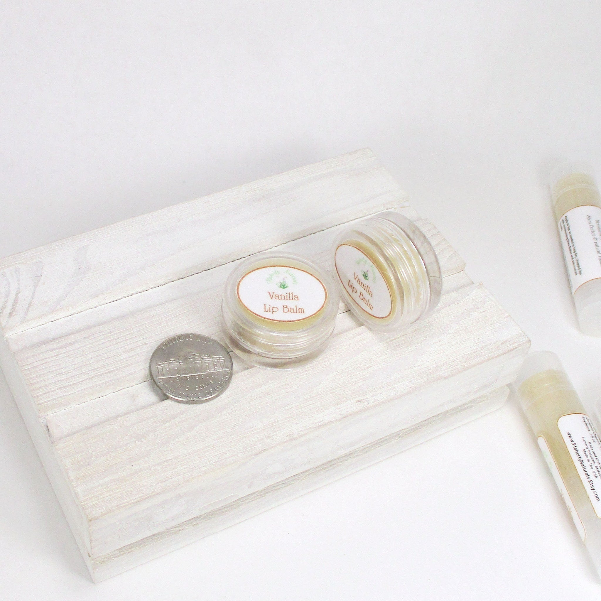 Lip balm in clear plastic containers with white labels, on a white wooden tray, with a coin