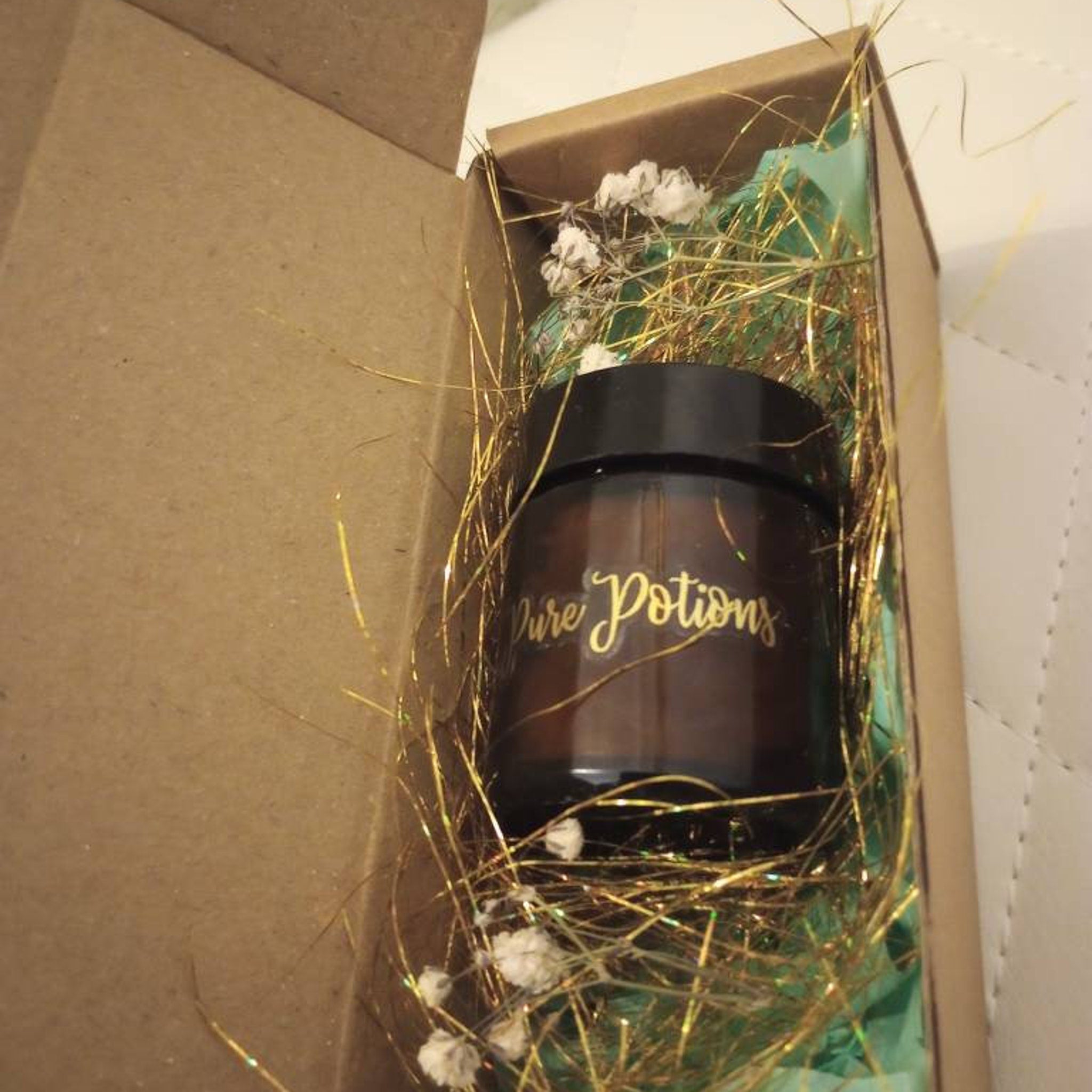 Brown jar with black lid, gold text "Pure Potions" in a brown box with gold filler and white flowers