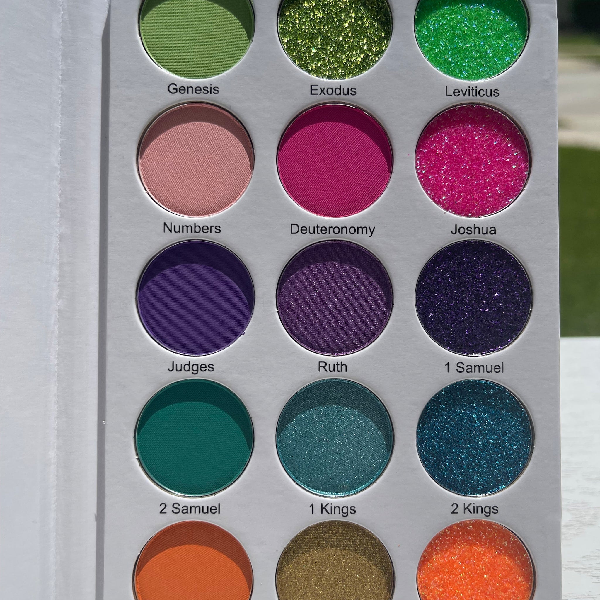 Eyeshadow palette with green, pink, purple, and teal shades, in matte and glitter finishes, with white packaging