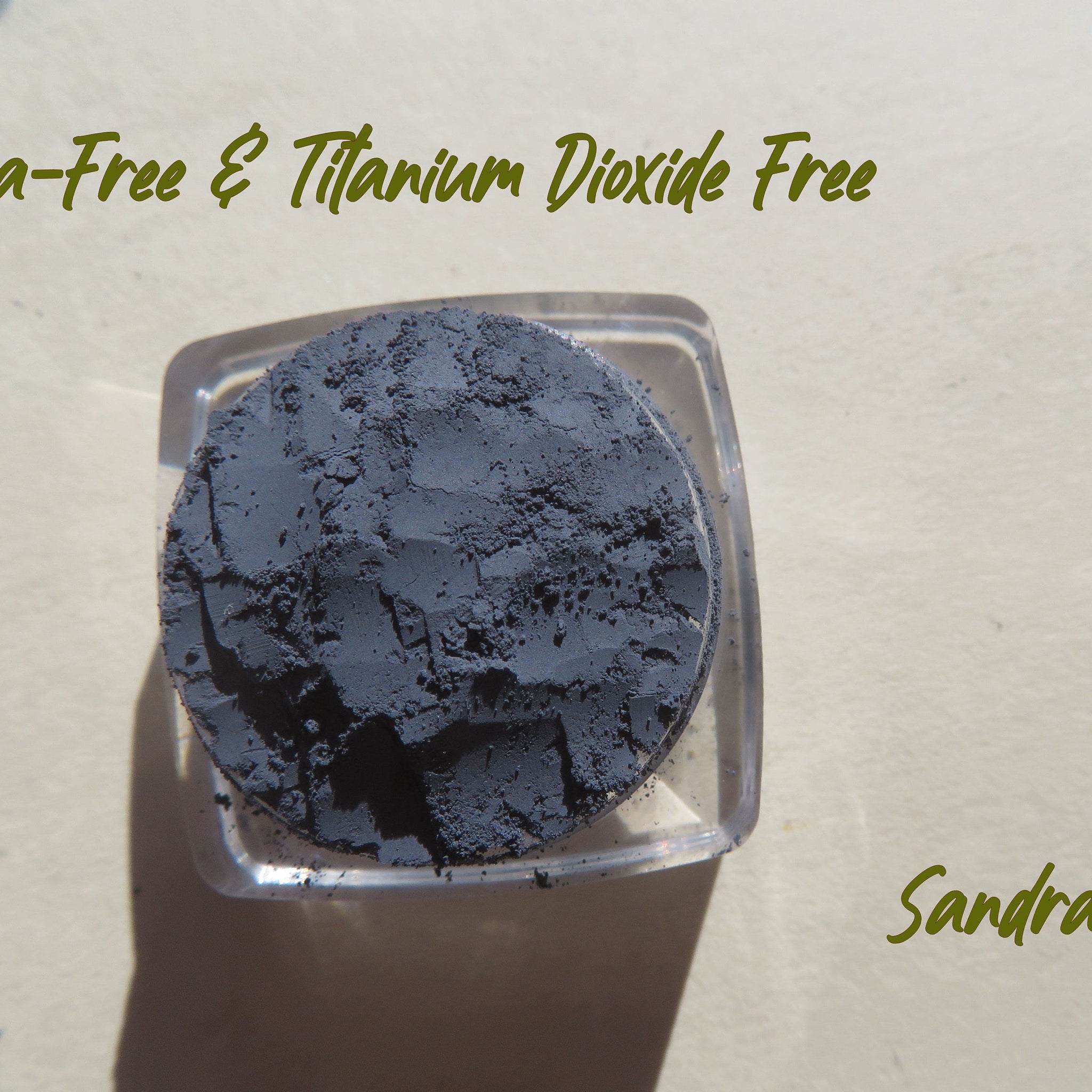 Powdered dark gray product in clear square container, textured surface, Sandra text