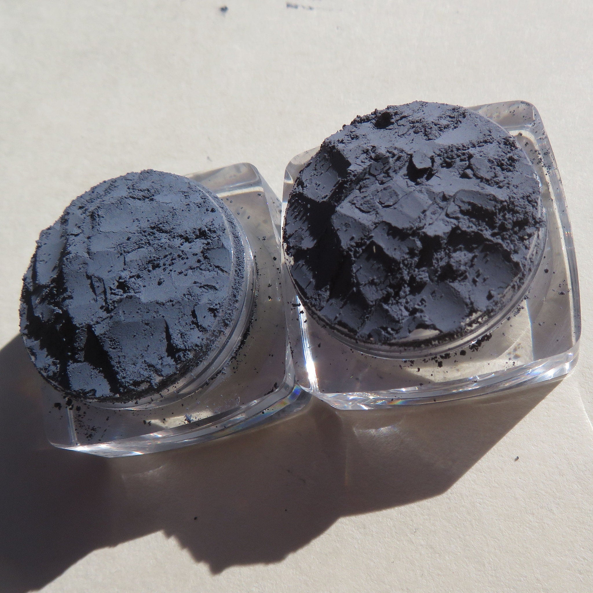 Powder eyeshadow, dark grey, textured, in clear square containers on light surface