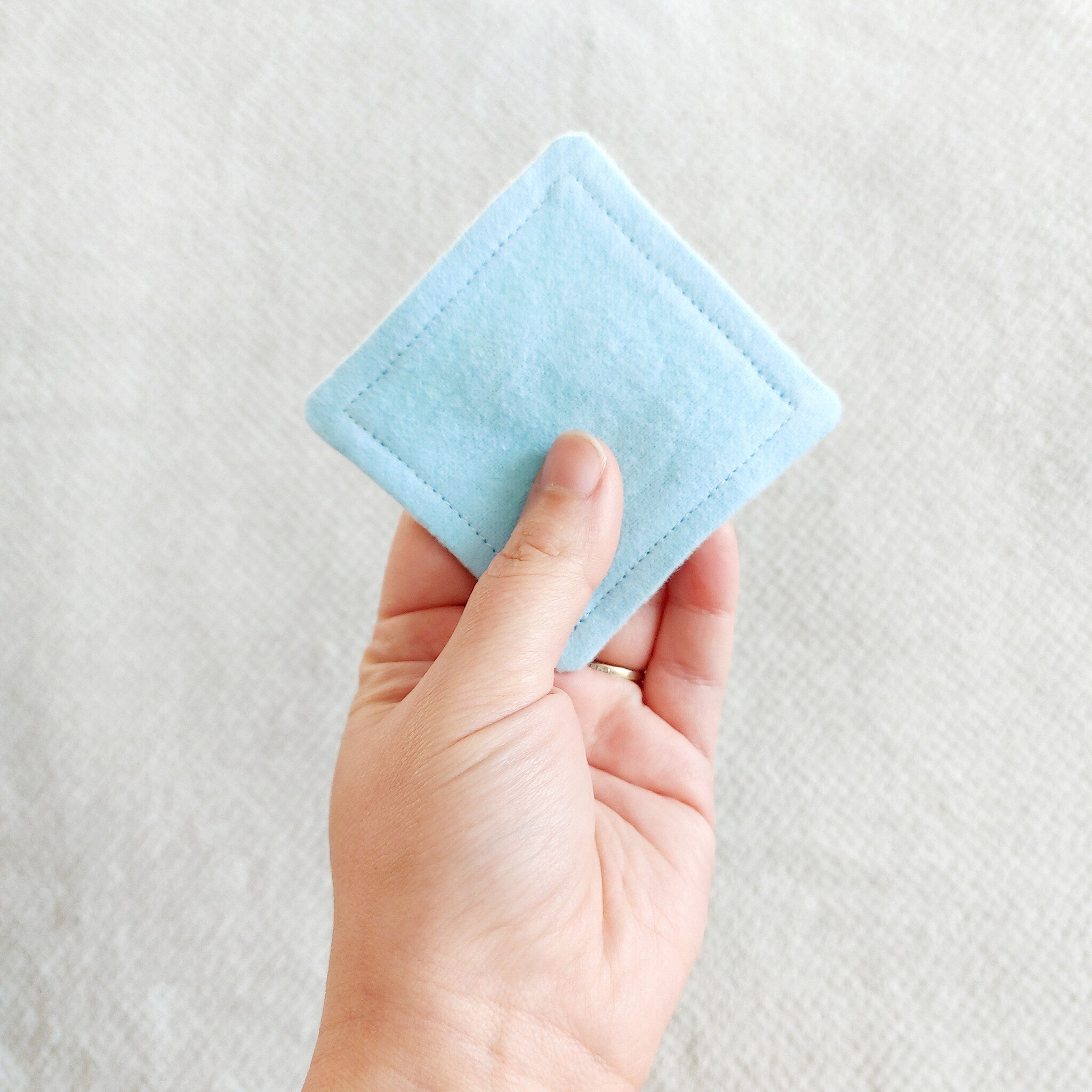 Reusable makeup remover pad, light blue felt, stitched edge, held in hand