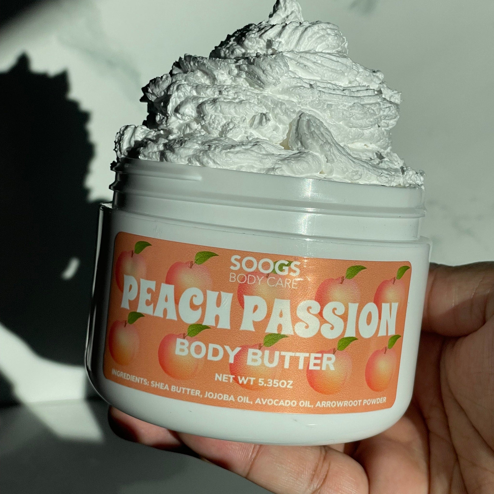 Body butter jar with white cream, peach label, and hand holding it SOOGS brand