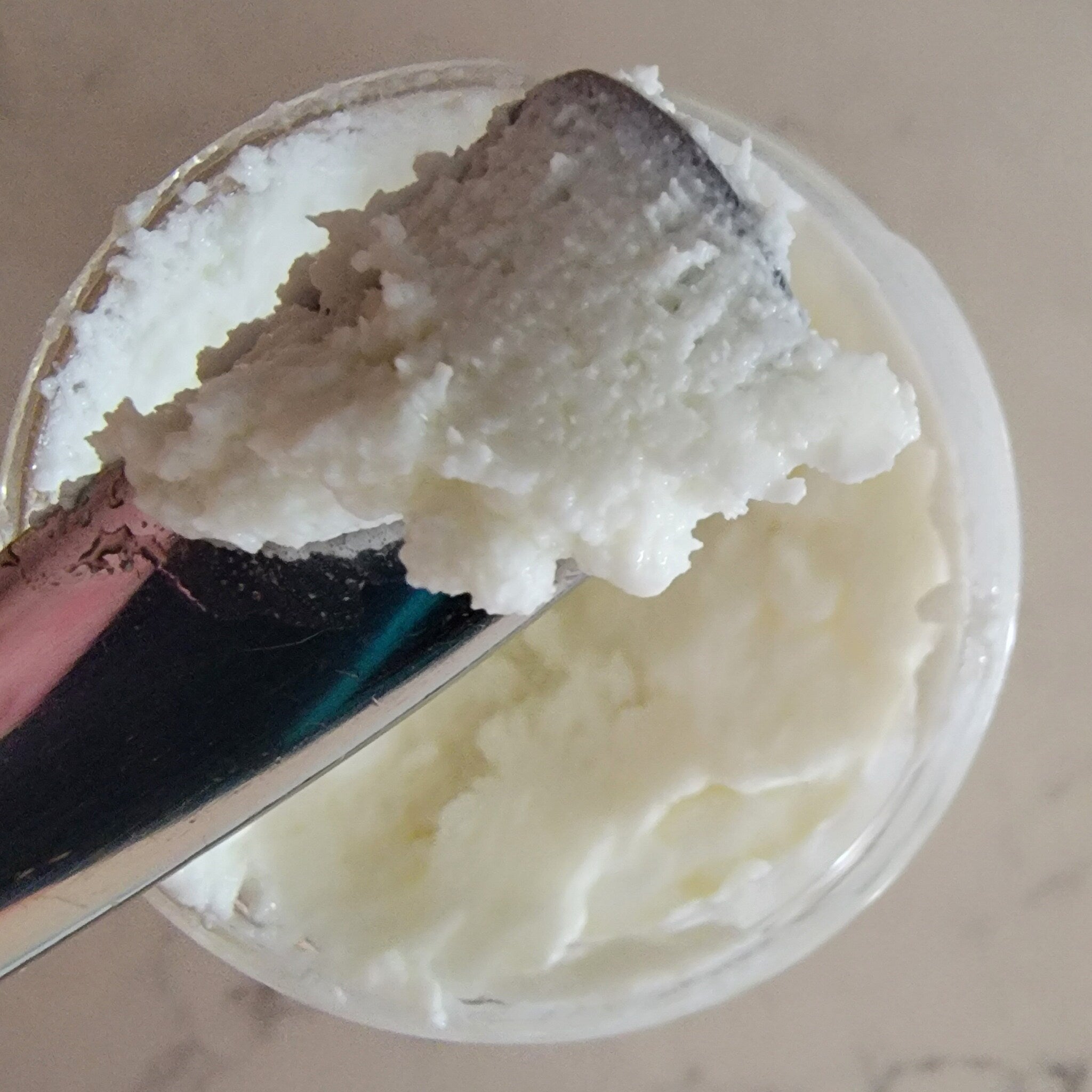 White cream with a metal utensil, in a clear container, on a light surface