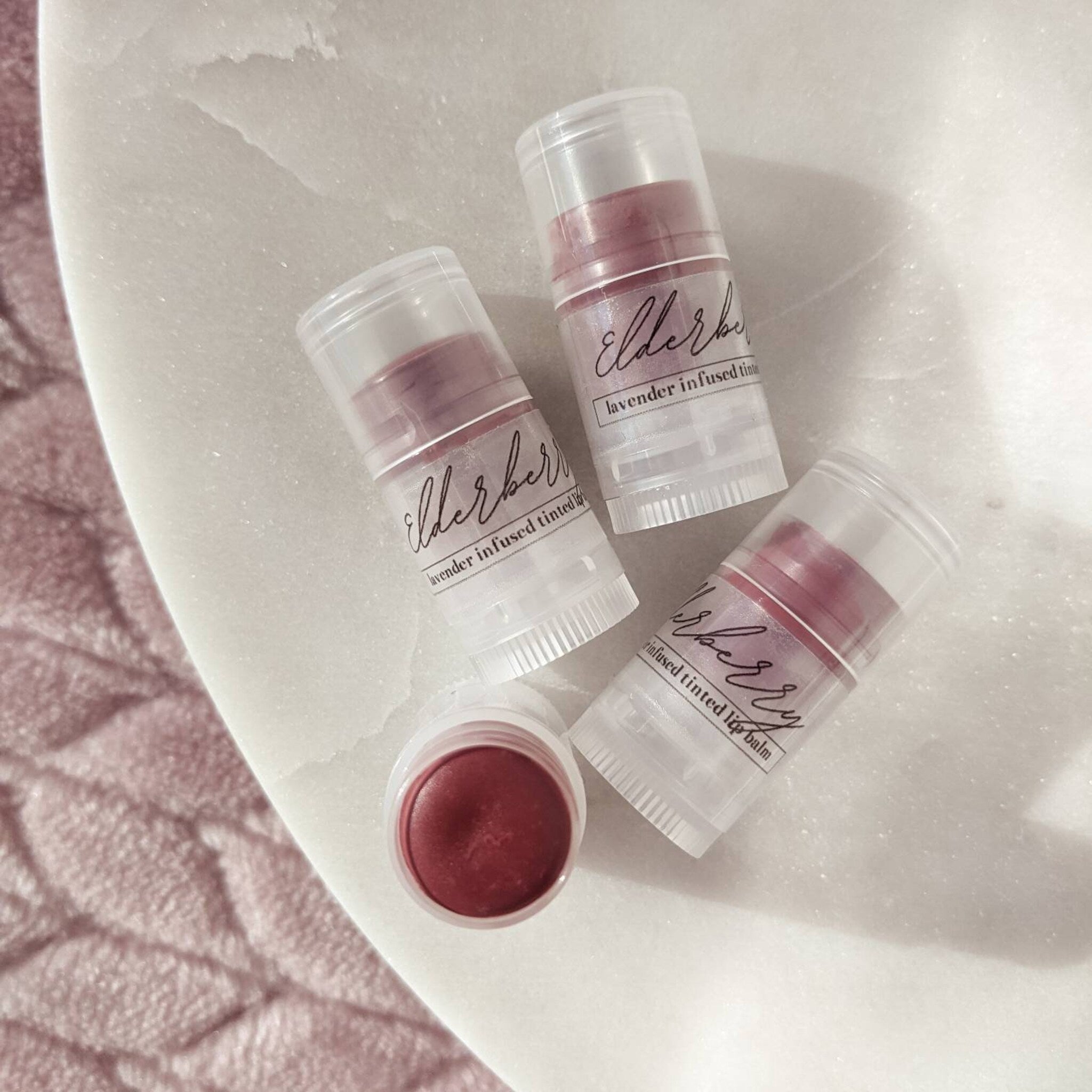 Tinted lip balm in clear tubes with burgundy balm, "elderberry" label, on white surface