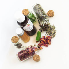 Bottles with corks and herbs, glass jars with dried rose petals, and a green soap bar on white background