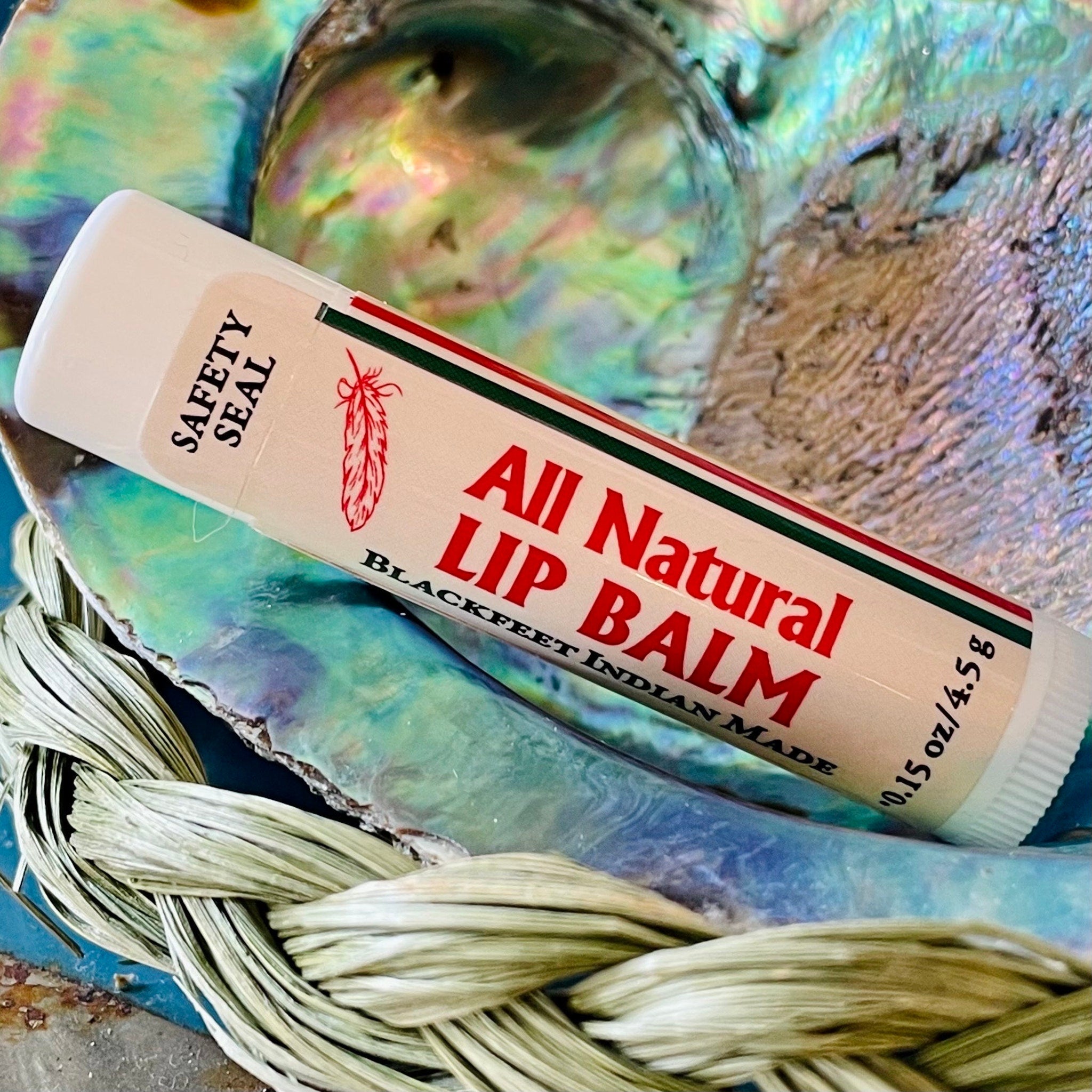 Lip balm tube with white, red, and green stripes, "All Natural Lip Balm" text, and feather logo on a shell background