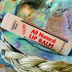 Lip balm tube with white, red, and green stripes, "All Natural Lip Balm" text, and feather logo on a shell background