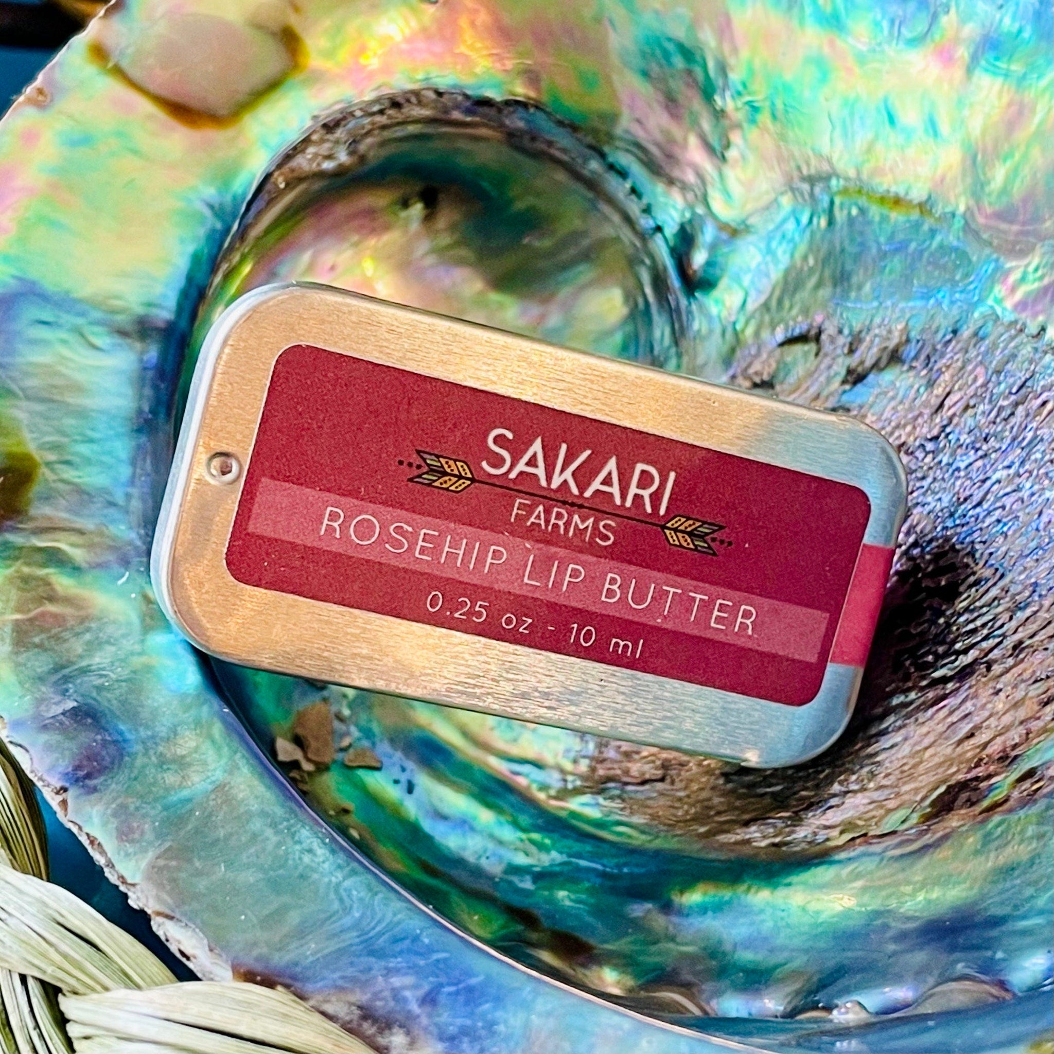 Lip butter tin with red label, gold trim, and Sakari Farms brand name, in a colorful shell