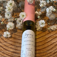 Lash oil bottle with pink cap, clear glass, and white label, surrounded by white flowers on wood