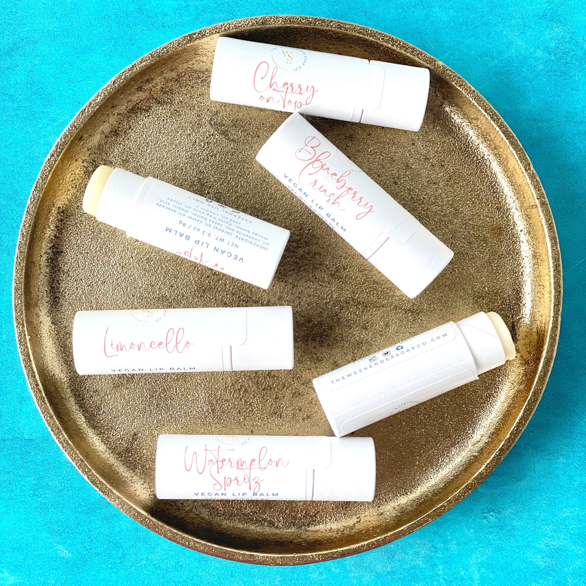 Vegan lip balms in white tubes with pink text on a gold tray on a blue surface
