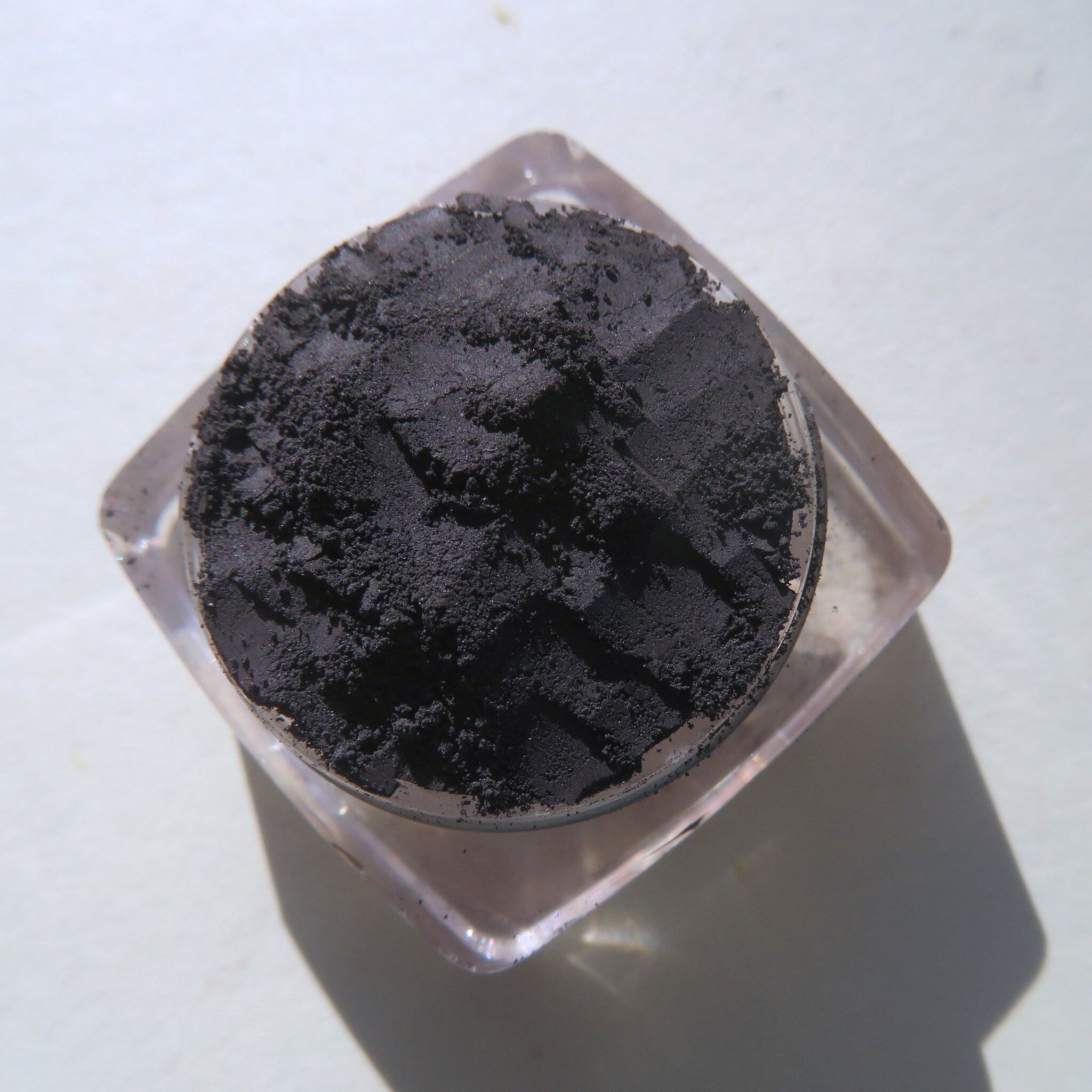 Black powder eyeshadow in clear plastic container, textured dark pigment, angled view