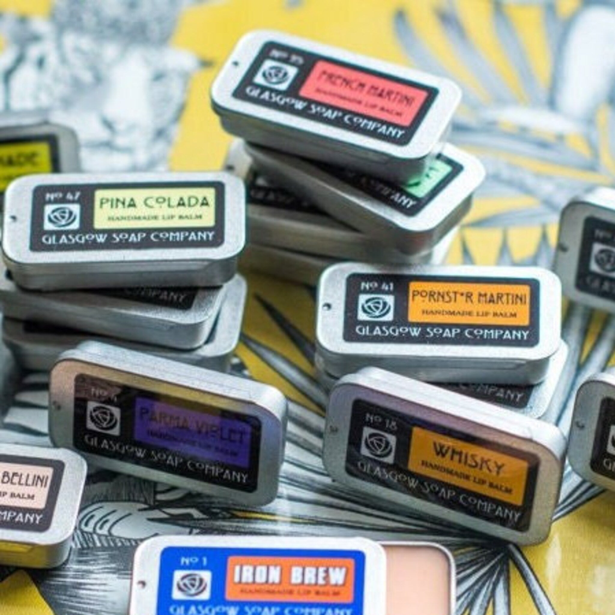 Lip balm tins with various color labels and text, stacked on yellow surface, Glasgow Soap Company visible